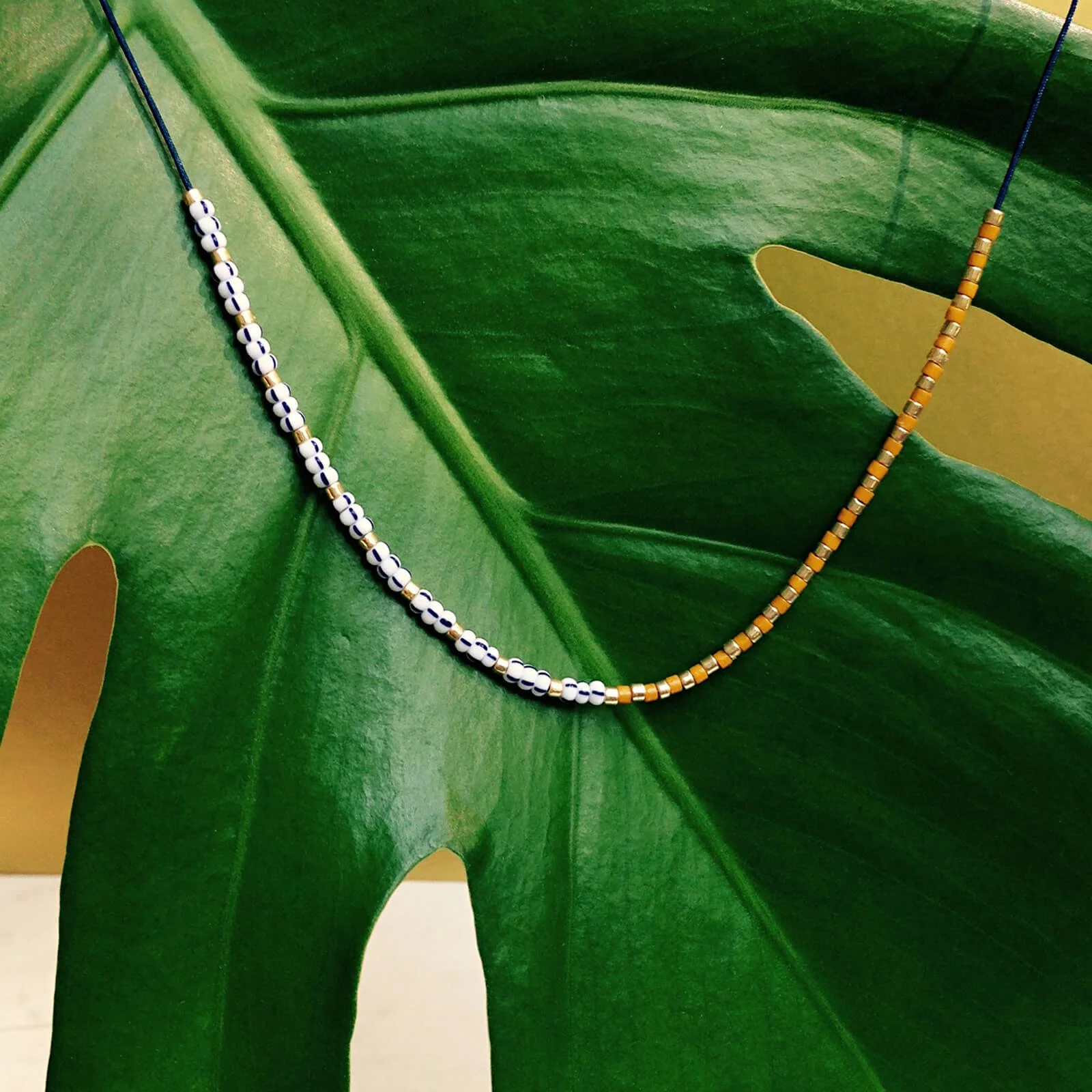 Elegante Halskette auf dunkelblauem Nylonfaden – eine Hälfte mit dunkelblau-weiß gestreiften Glasperlen, die andere im Streifenlook von honig- und goldfarbenen Perlen – auf einem großen Blatt