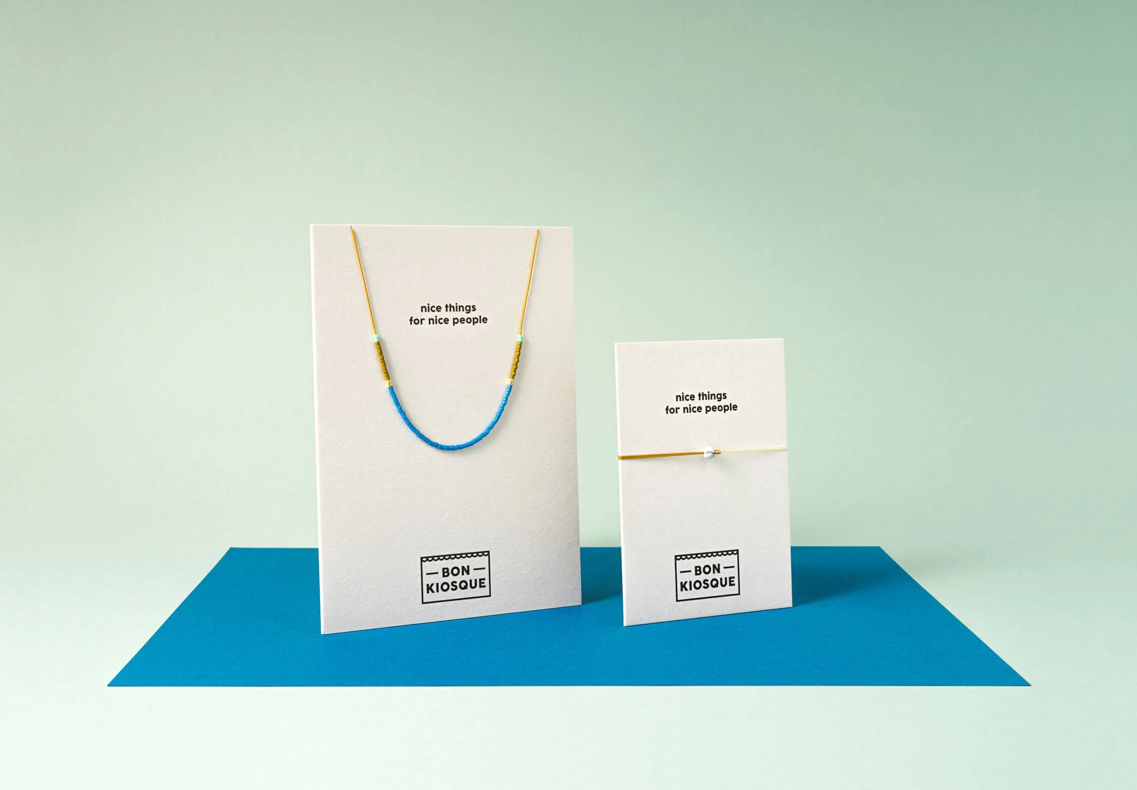 BON KIOSQUE Schmuck auf weißen Produktkarten vor mintgrünem Hintergrund: eine Halskette aus blauen und olivefarbenen Glasperlen auf goldenem Nylonfaden sowie ein Fullmoon-Armband auf ockerfarbenem Nylonfaden mit Howlith-Stein