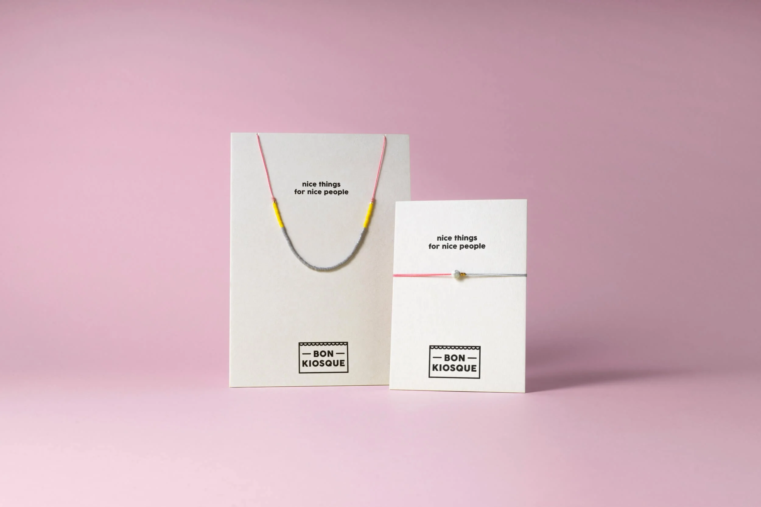 BON KIOSQUE Schmuck auf weißen Produktkarten vor rosafarbenem Hintergrund: eine Halskette aus grauen und gelben Glasperlen auf pinkem Nylonfaden sowie ein Fullmoon-Armband auf grauem und rosafarbenem Nylonfaden mit Howlith-Stein