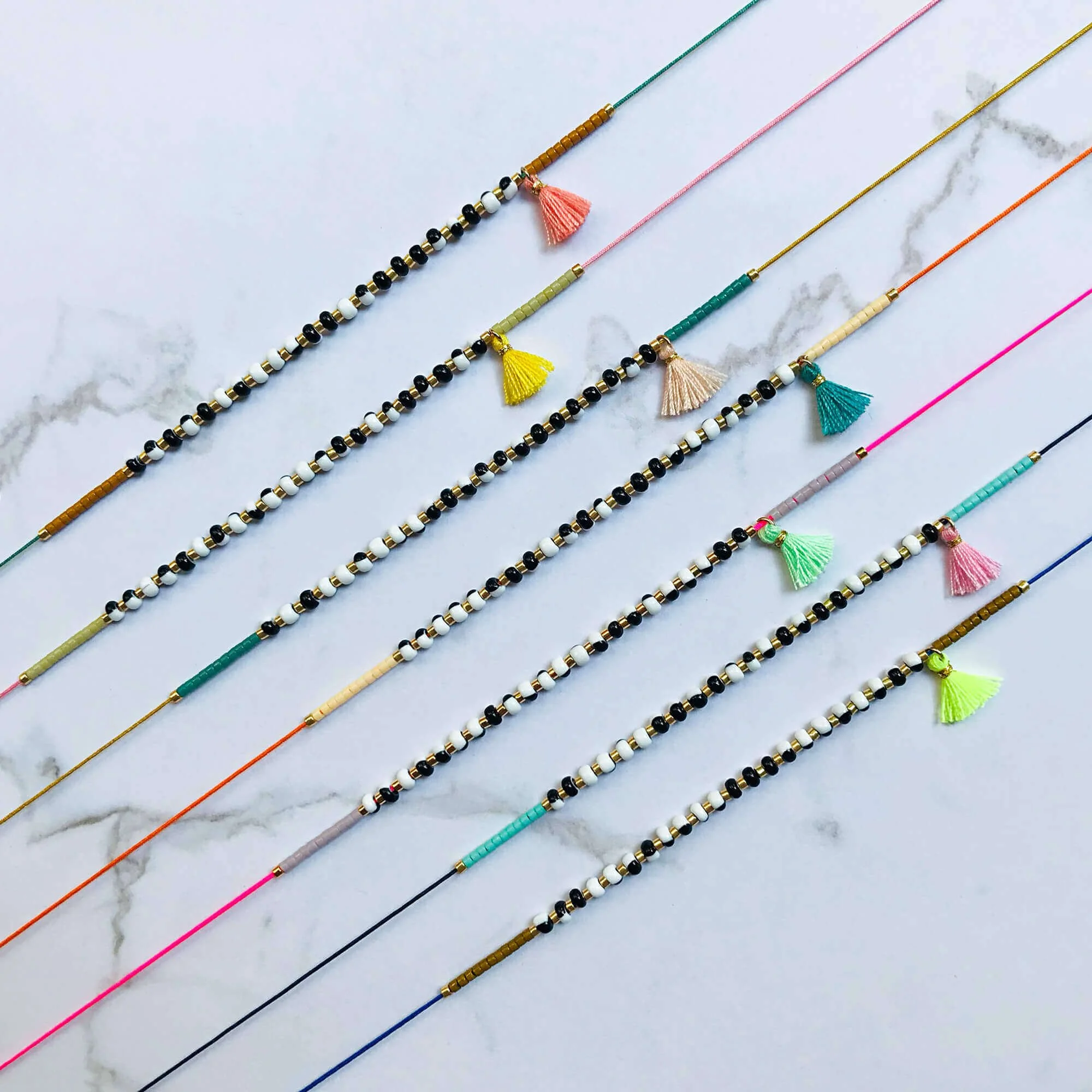 Alle 7 BON KIOSQUE Halsketten der Pompon Kollektion nebeneinander auf Marmor – schwarzweiße Glasperlen mit bunten Pompons in Korall, Gelb, Apricot, Petrol, Mint, Rosa und Neongelb