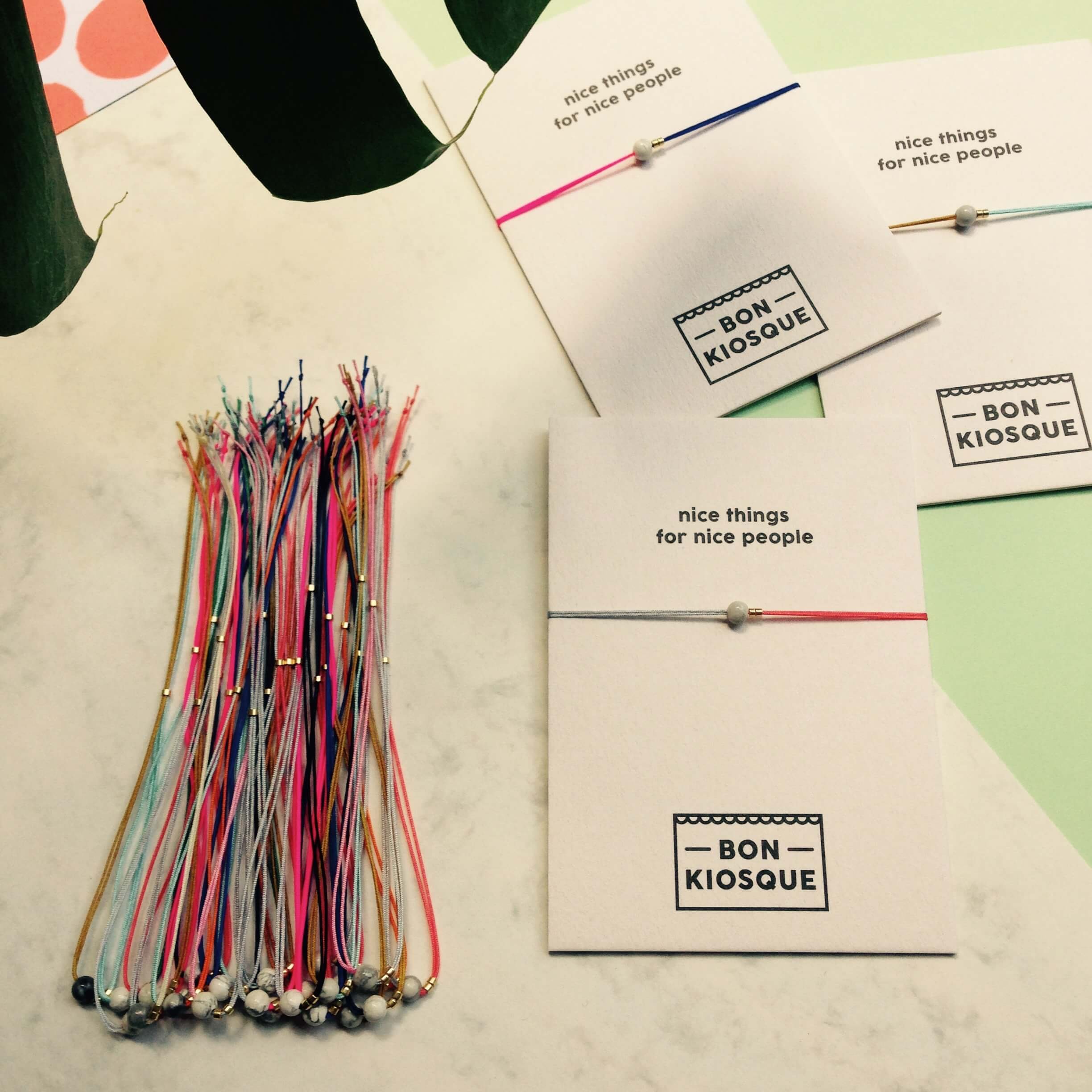 Mehrere BON KIOSQUE Fullmoon-Armbändchen auf weißen Produktkarten mit Aufschrift nice things for nice people sowie ein Bündel Armbändchen in verschiedenen Farben auf Marmor, umgeben von grafischen Papieren und einem Monstera-Blatt