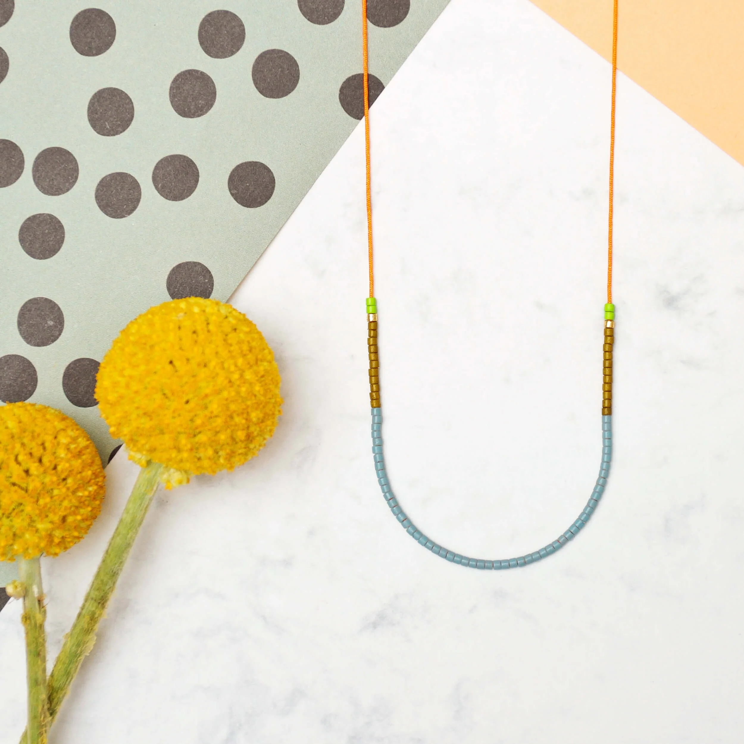 Filigrane Halskette aus kleinen Glasperlen in Eukalyptus, Olive und Limette auf orangenem Nylonfaden, auf Marmor mit gelben Trockenblumen und gepunktetem Papier arrangiert
