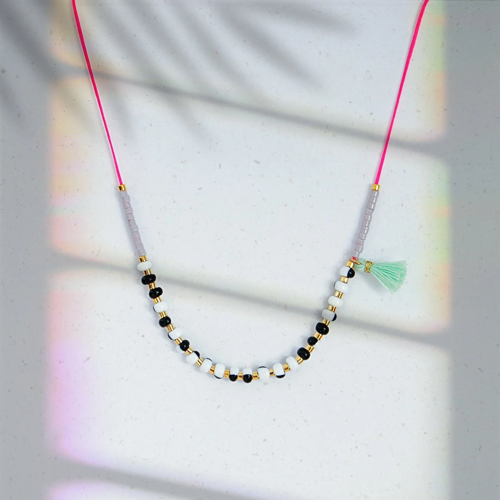 Zarte Halskette aus schwarzweißen, gold- und grauen Glasperlen auf pinkem Faden mit mintfarbenem Pompon, vor hellem Hintergrund mit Palmschatten und irisierendem Lichtreflex