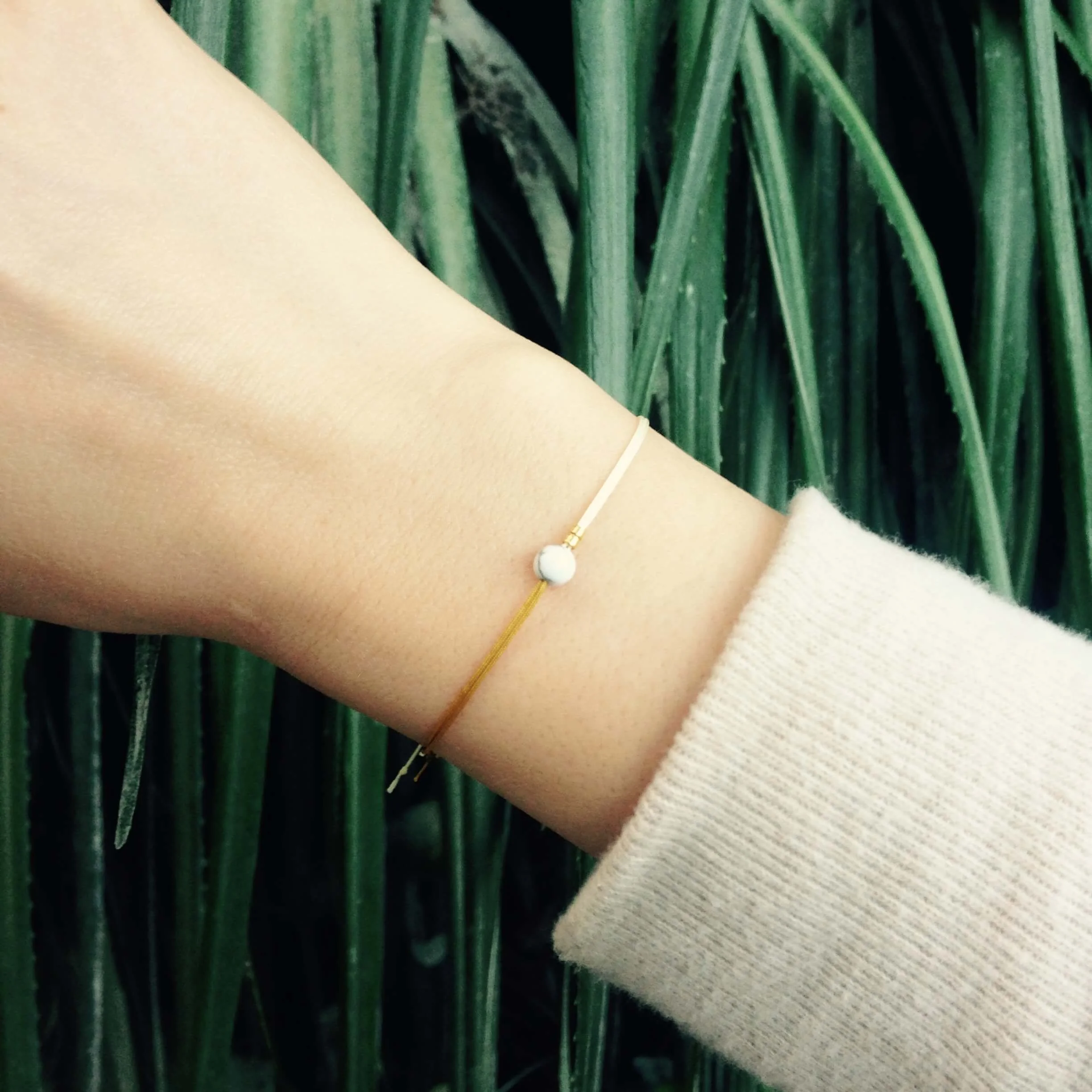 Zartes Fullmoon-Armband in Ocker und Creme mit Howlith-Stein am Handgelenk getragen, vor grünem Pflanzenhintergrund
