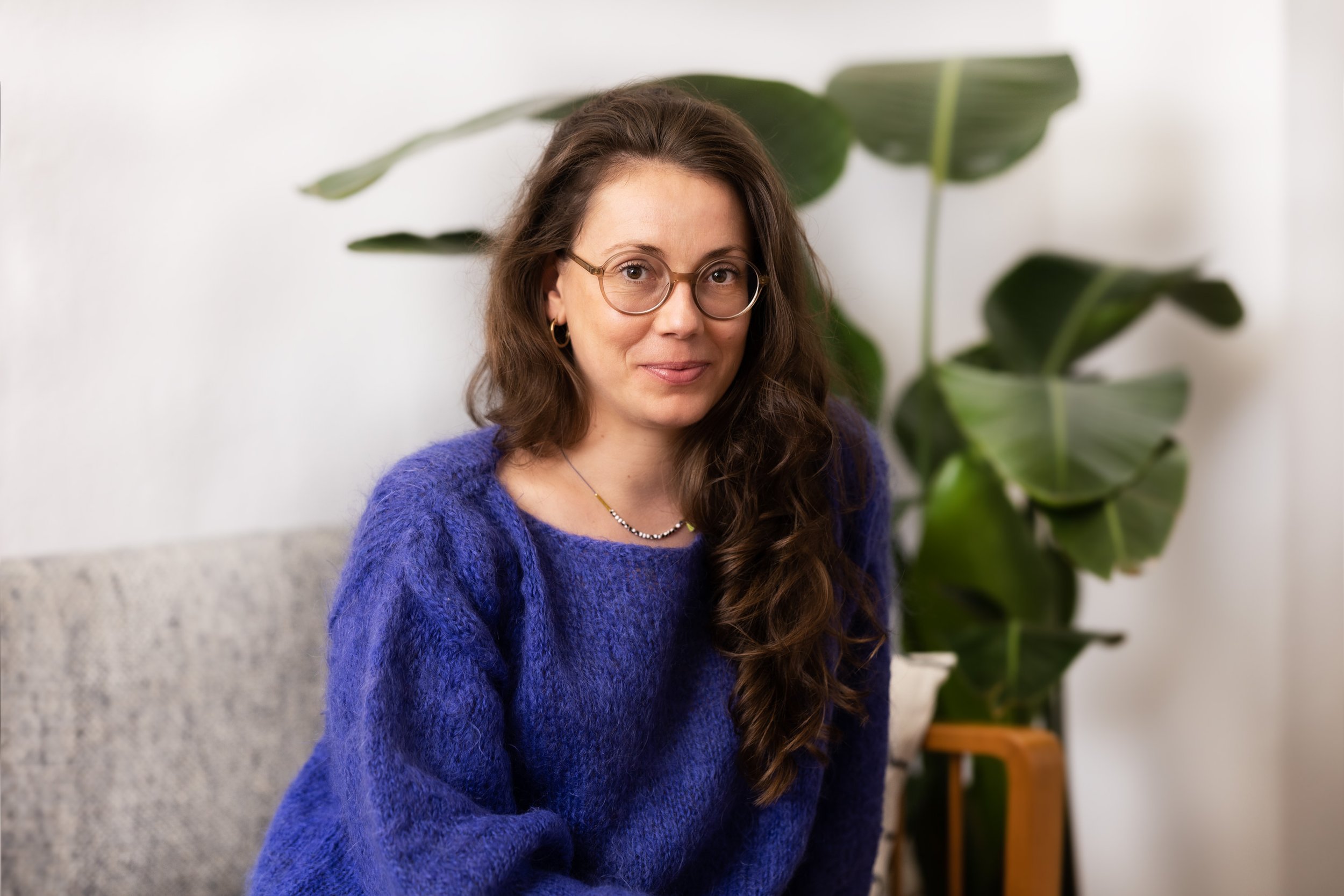 Porträt der BON KIOSQUE Gründerin: eine Frau mit langen braunen Haaren, runder Brille und blauem Mohairpullover, trägt eine zarte Glasperlen-Halskette, sitzt vor einer Monstera-Pflanze