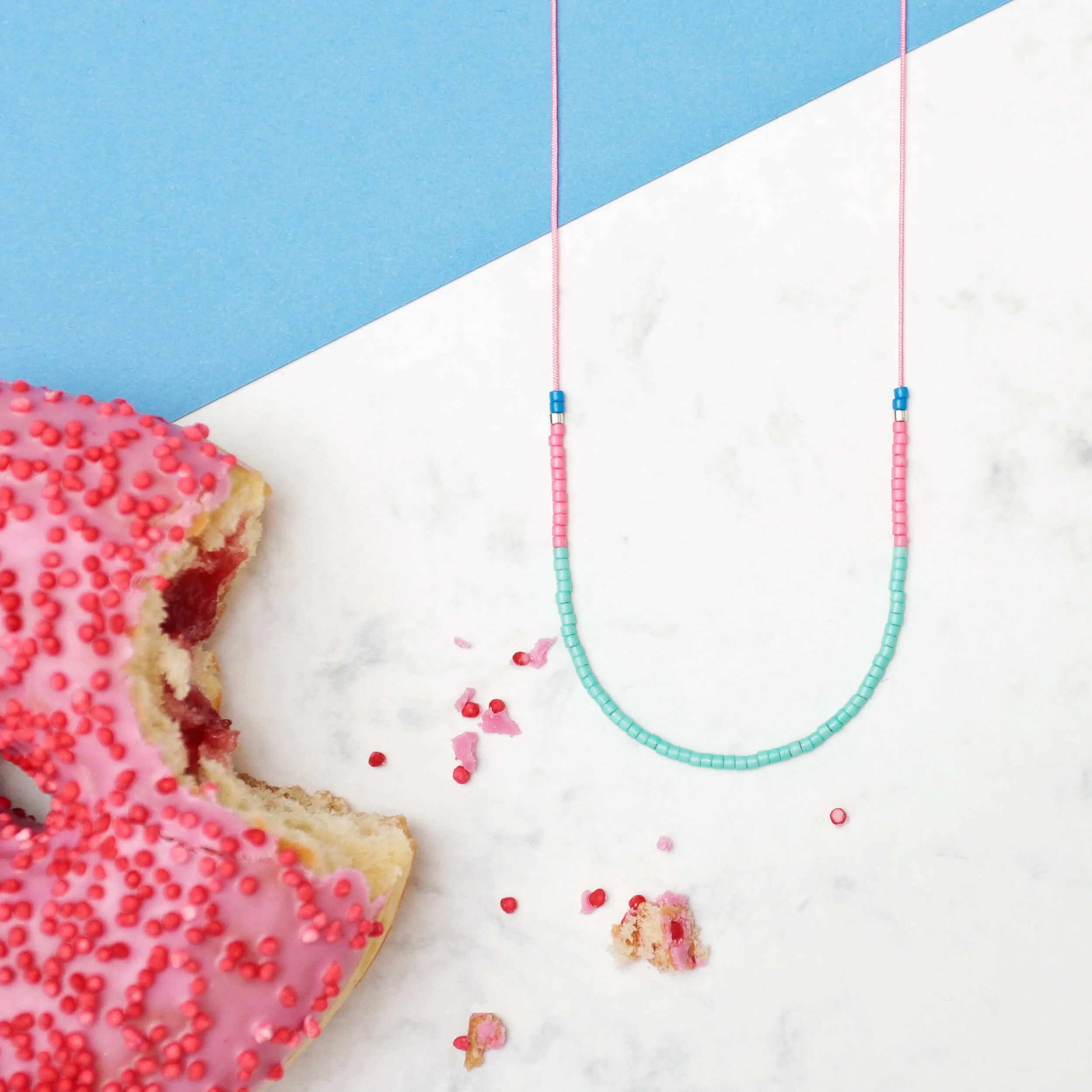 Filigrane Halskette aus kleinen Glasperlen in Mint und Pink auf rosafarbenem Nylonfaden, auf Marmor mit einem angebissenen rosa Donut arrangiert