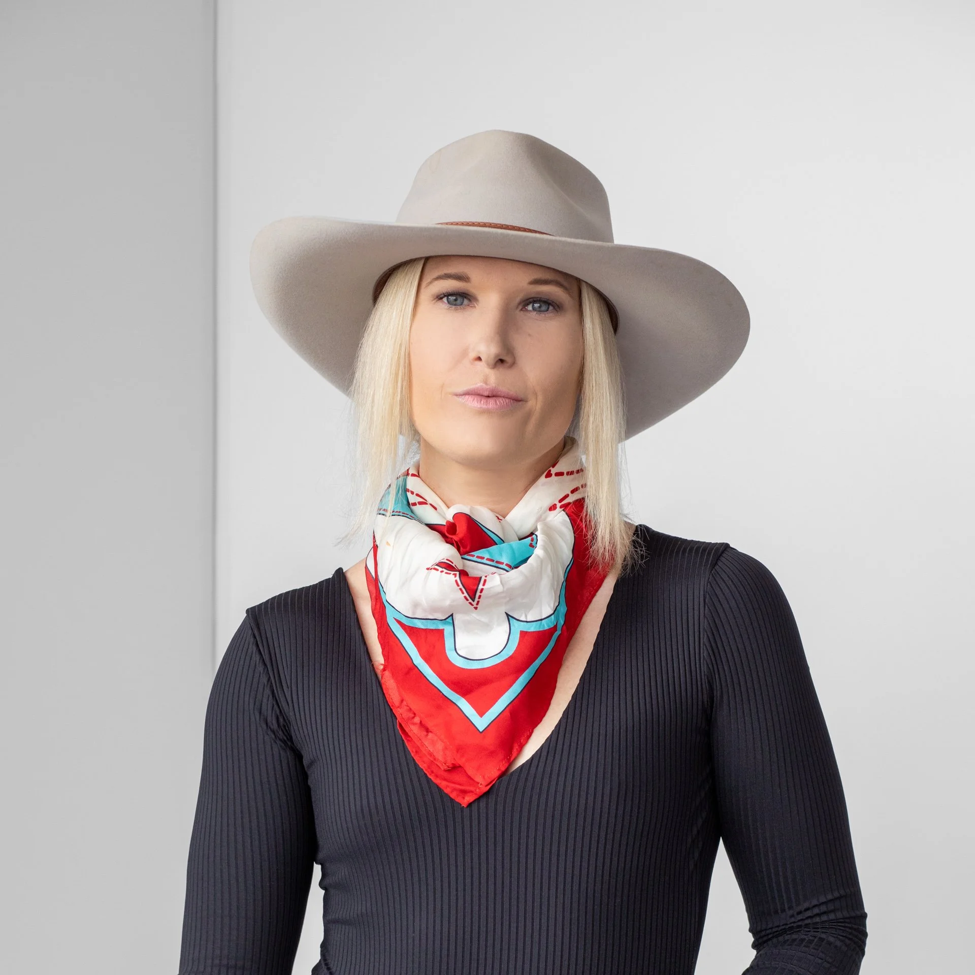 An indoor headshot of Kelcee. She wears a black long sleeve, with a colorful bandana and cream cowboy hat as she stares cooly at the camera.