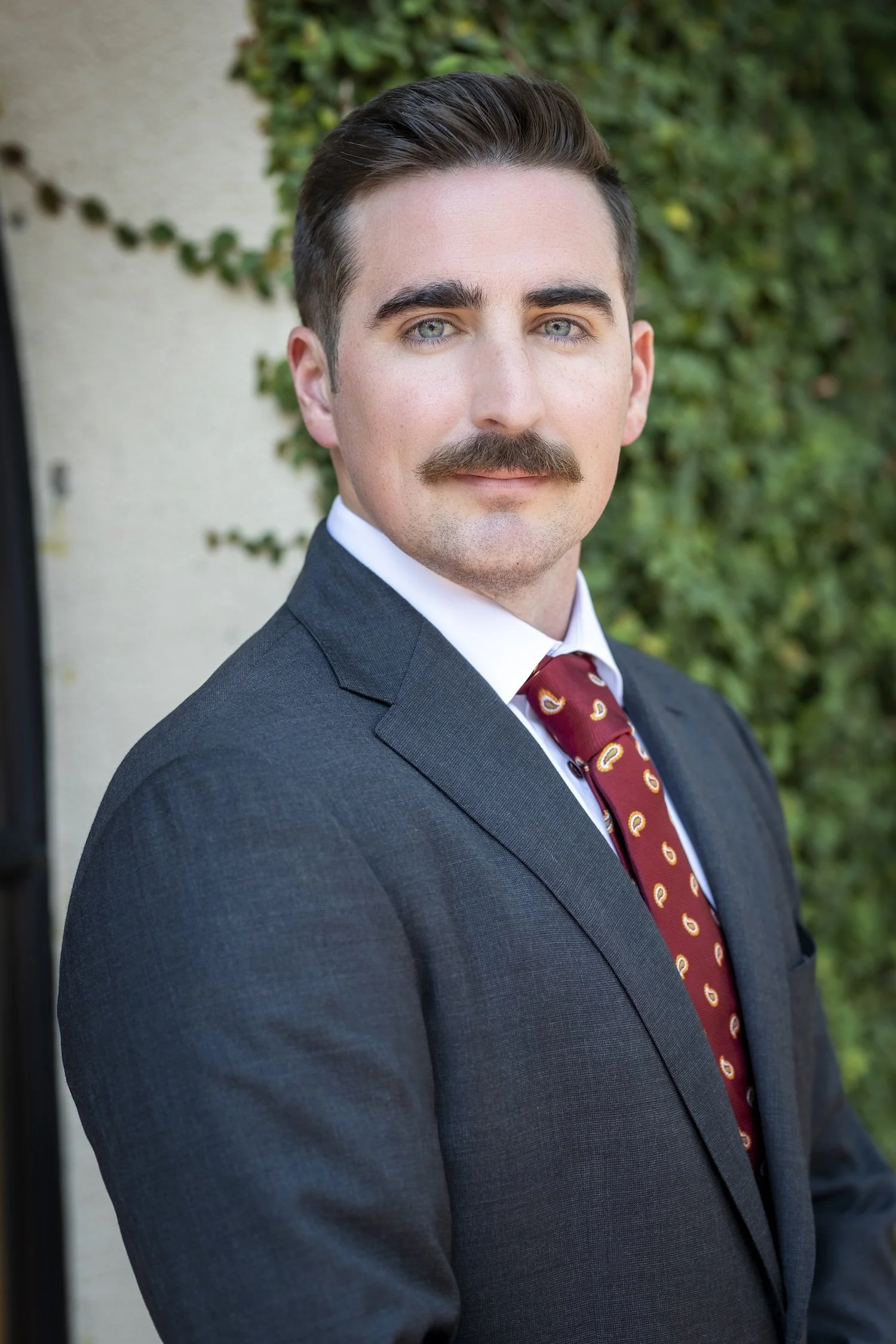 An outdoor headshot of a white man. He wears a dark gray suit and a red paisley tie. He smiles softly at the camera. There's a white building with green ivy climbing the wall in the background.