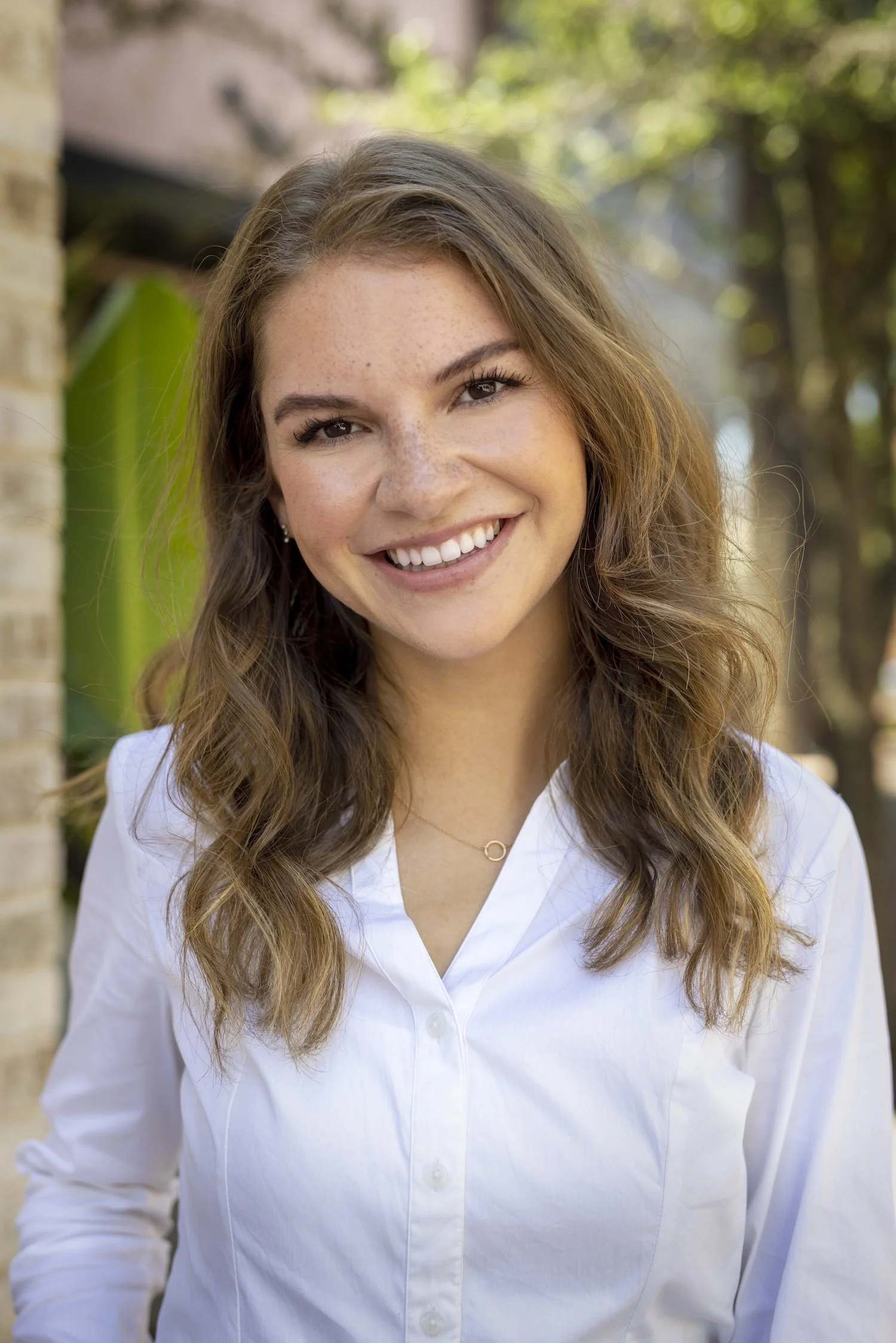 An outdoor headshot of a young white woman. She wears a white button down and smiles brightly at the camera. Tropical plants and a tan building can been seen peaking in from the background.