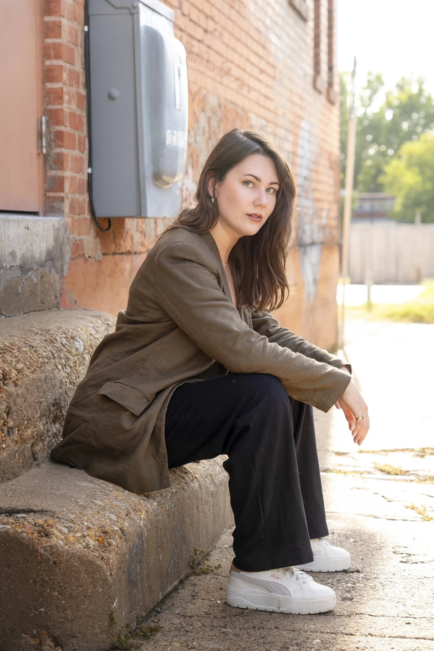 A side view of Megan as she looks at the camera confidently with a relaxed pose as she sits on a set of concrete steps. She wears a brown blazer, black slacks, and white sneakers.