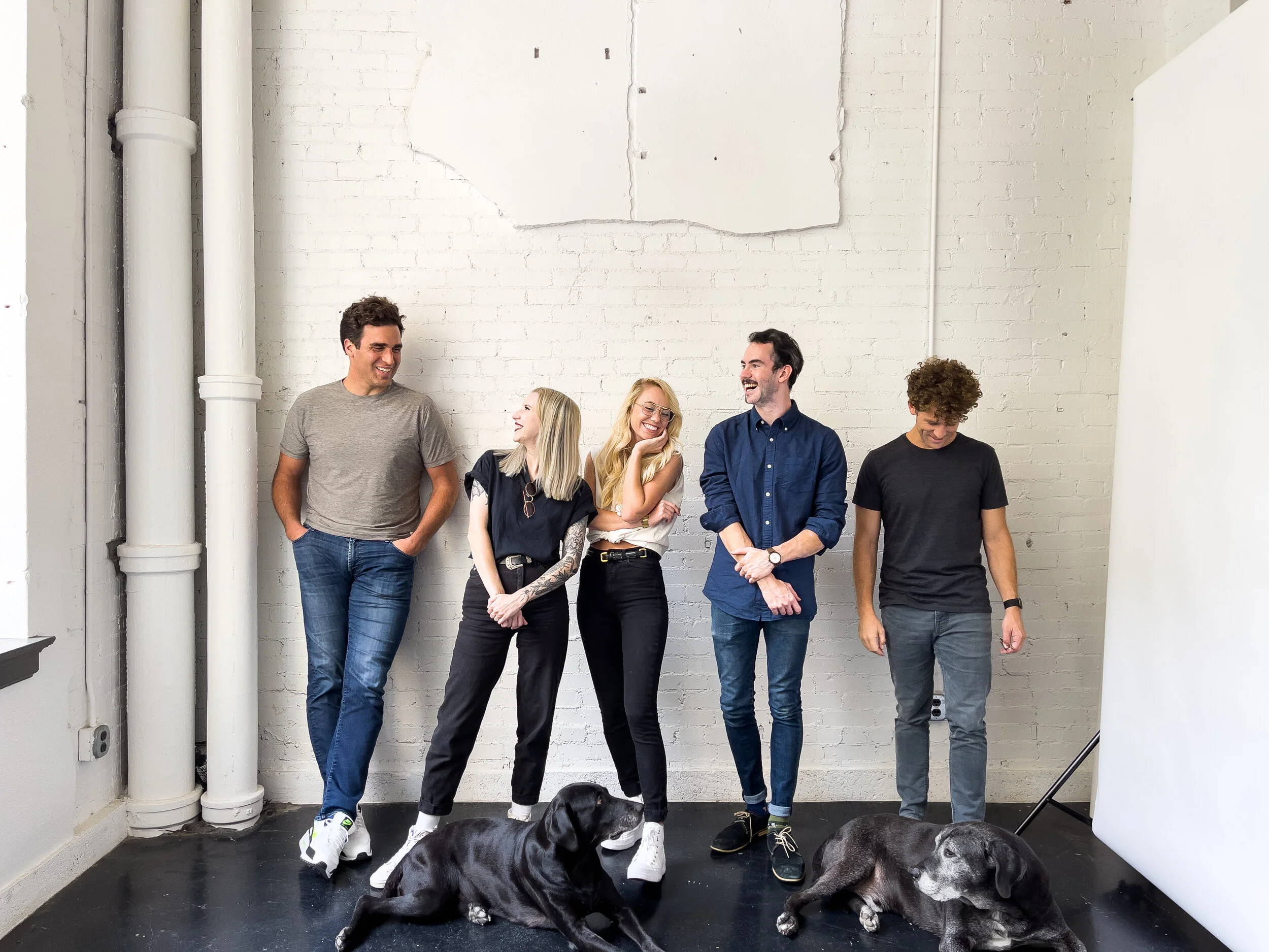 A team headshot of five Dallas professionals. They are dressed casually, with two dogs at their feet, as they stand laughing together.