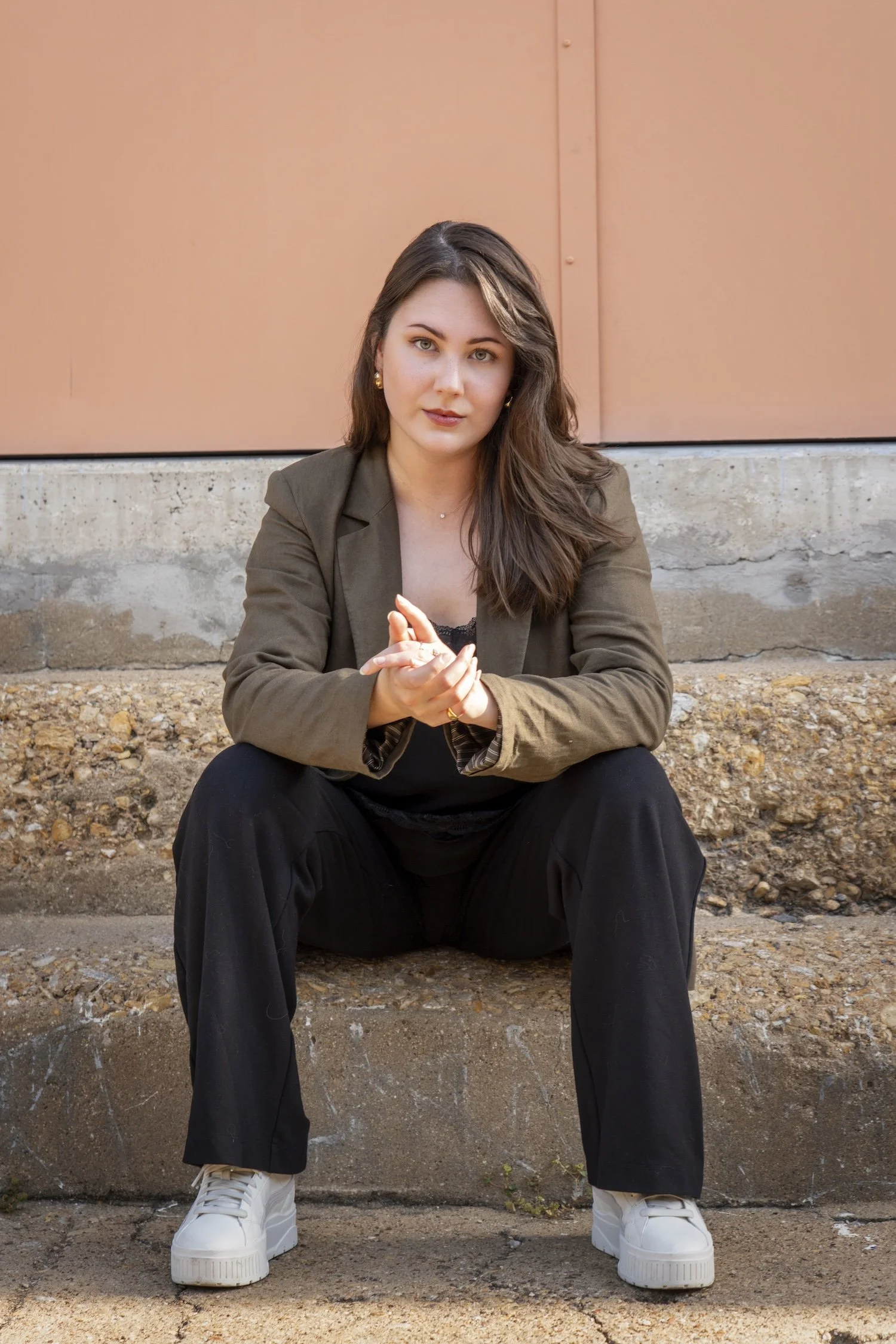 A front view of Megan as she looks at the camera confidently with a relaxed pose as she sits on a set of concrete steps. She wears a brown blazer, black slacks, and white sneakers.