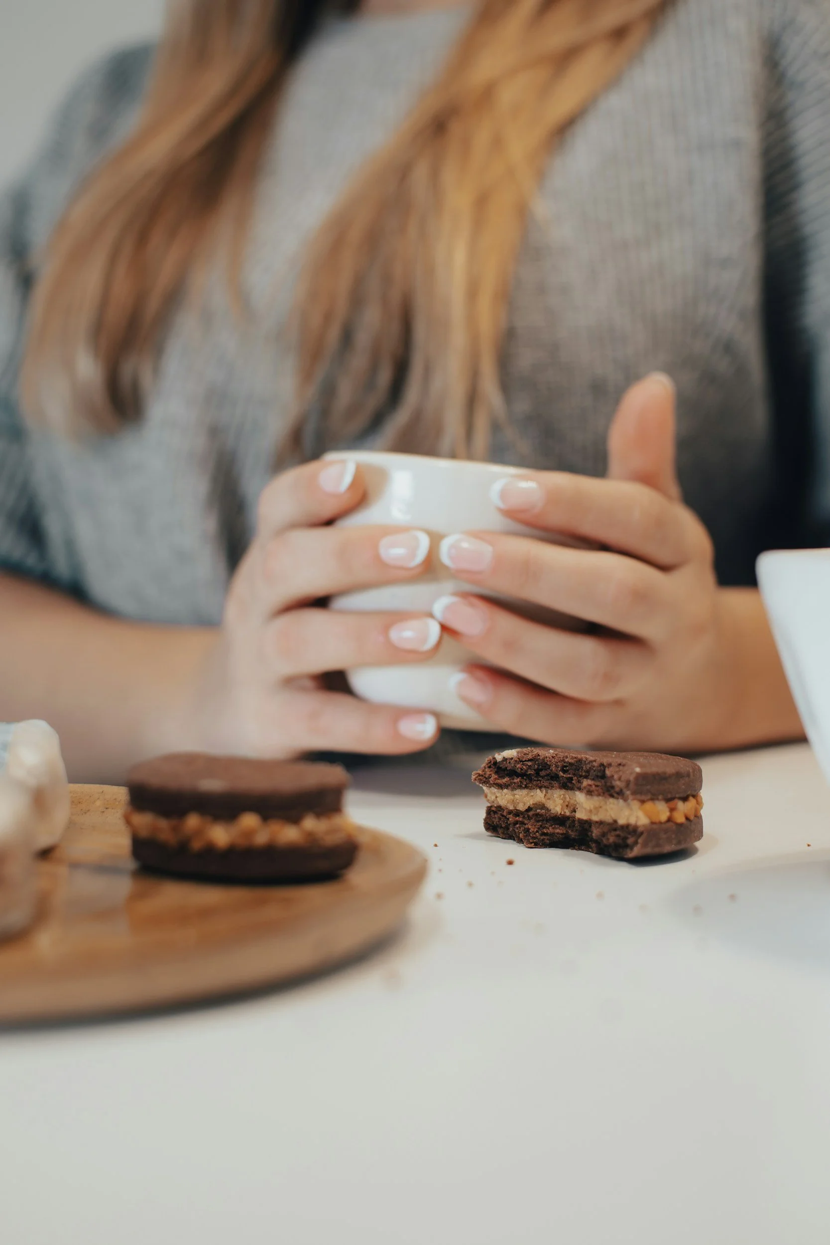 A chocolate biscuit broken in half in front of a midlife woman holding a white cup in both of her hands as she thinks about snacking to improve her energy.