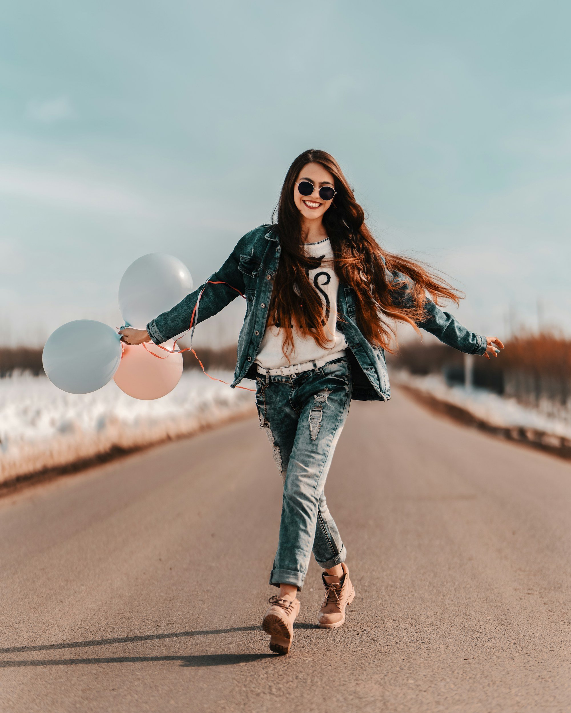 A woman with long brown hair walking on an open road with her arms open wide, carrying a bunch of pastel coloured balloons in one hand, smiling as she feels confident in who she is.