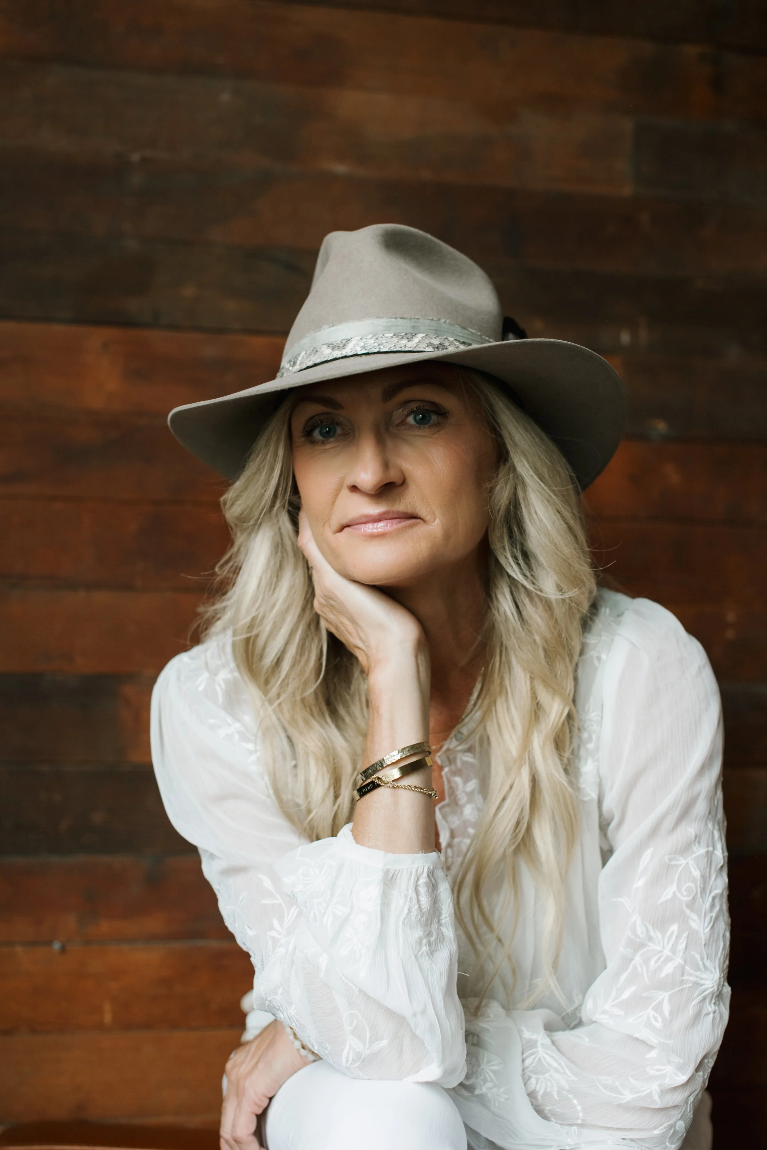 TIna LeAnn has long wavy blonde hair and wears a light beige hat, resting her chin onto one hand with three gold bracelets visible on her wrist as she shares why self-talk is so important to your health in menopause.