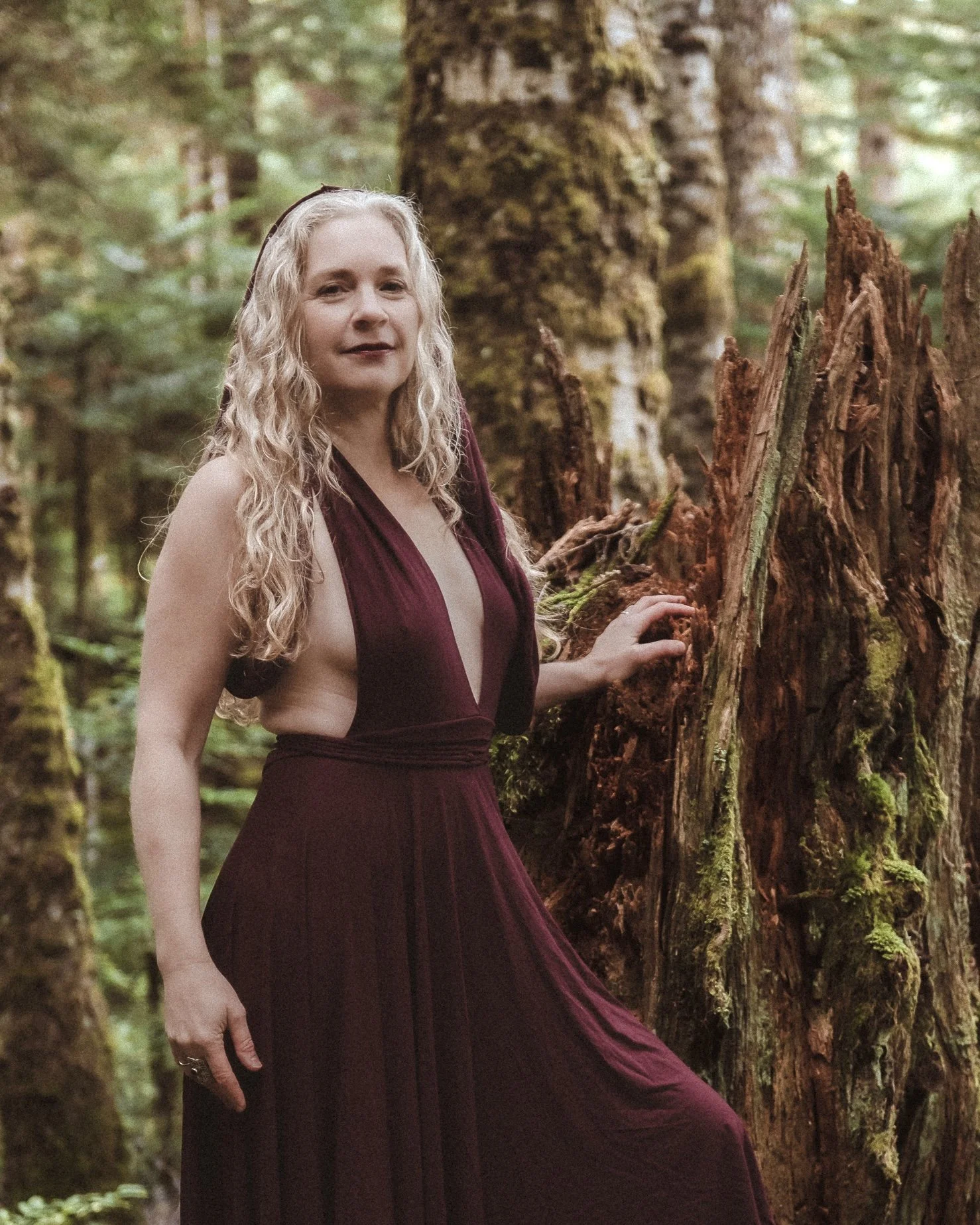 Carla Wainwright stands with one hand resting on the trunk of a broken tree, wearing a plum coloured backless dress, with light blonde curly hair hanging over her right shoulder.