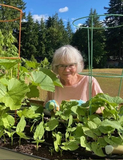 Jan Soloy standing in her garden wearing a pink top as she stands in front of her green plants holding a garden tool and wearing gardening gloves.