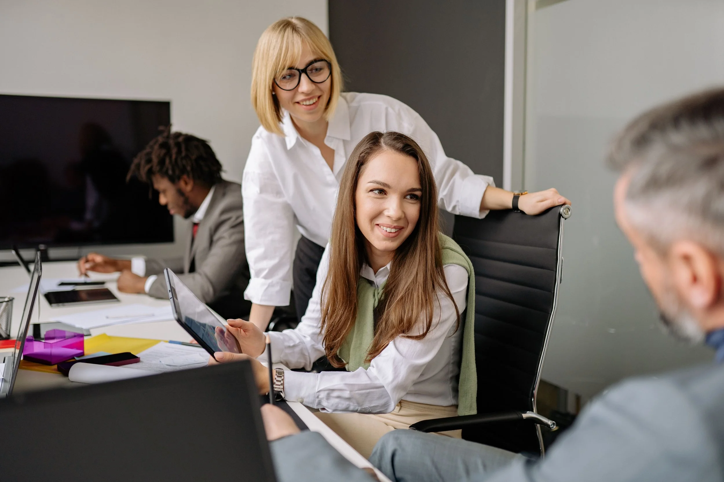 A team of two women and two men in a workplace setting, with both women smiling as they look towards one of the men who is just out of focus.