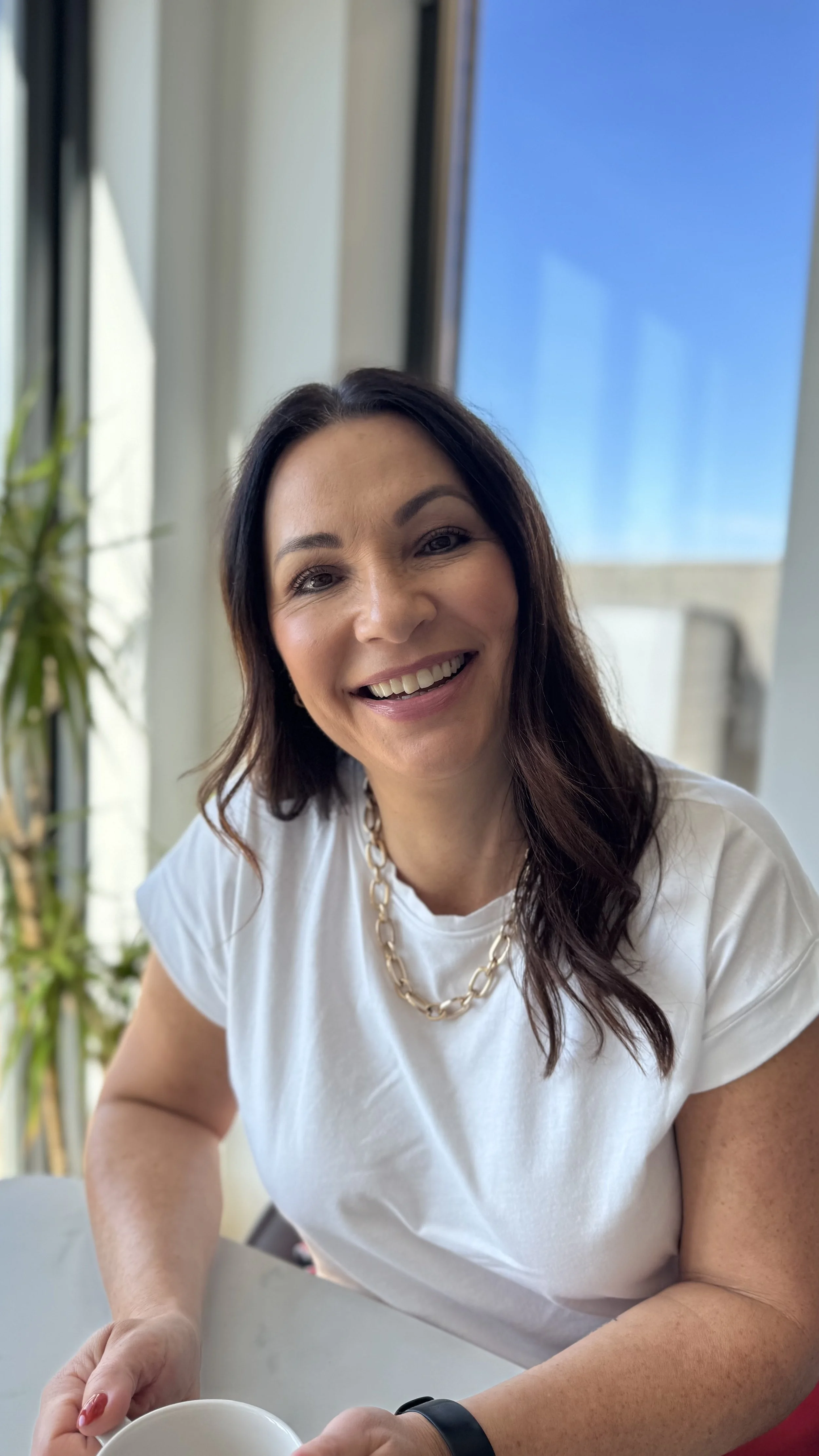 Nicola Farndell the host of the Women's Health Unwrapped podcast smiles wearing a chunky gold necklace and white t-shirt with her long brown wavy hair hanging over one shoulder.
