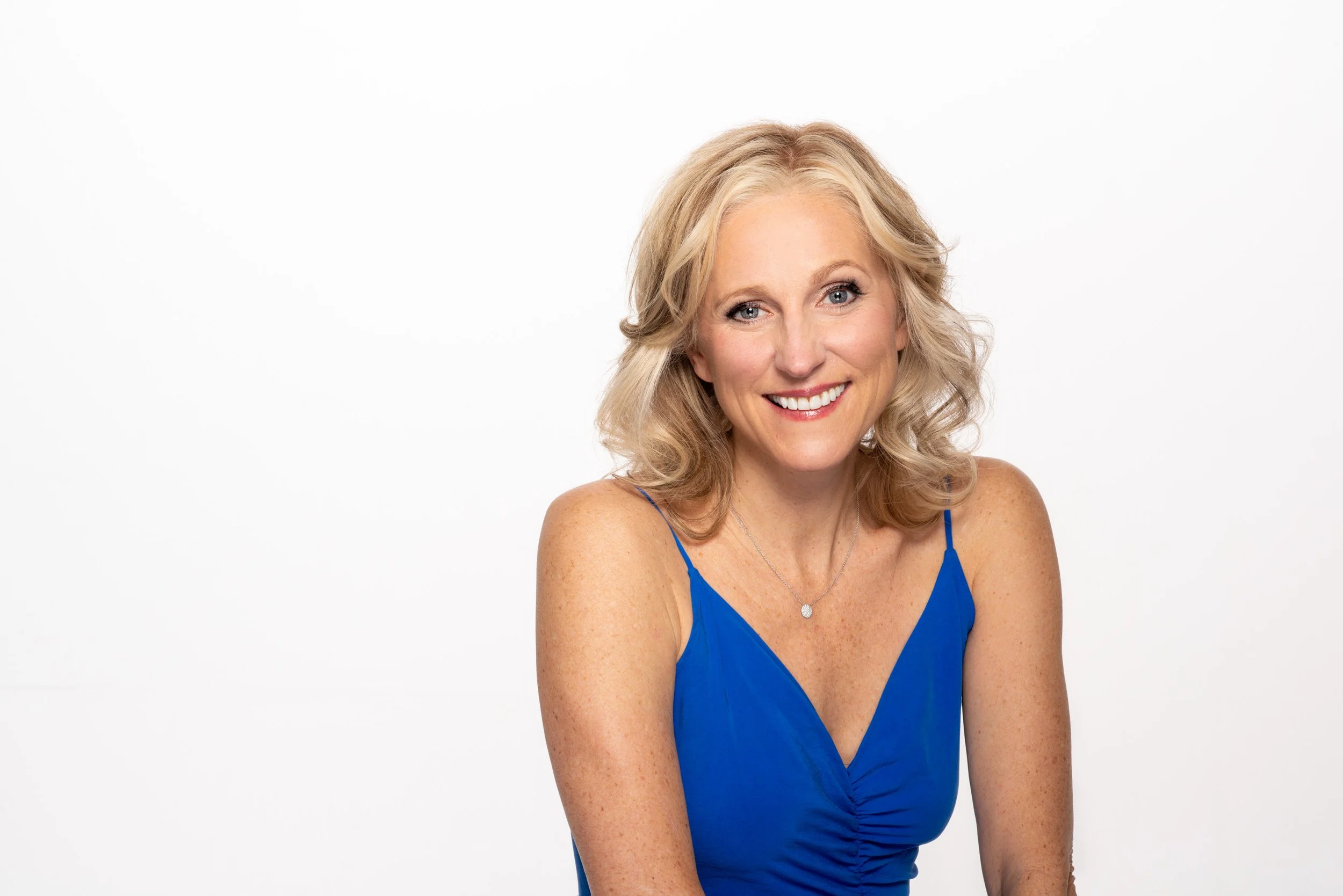 Tricia Nelson wears a bright blue dress with a gold necklace and blonde hair, smiling as she shares insights to help midlife women break free from emotional eating.