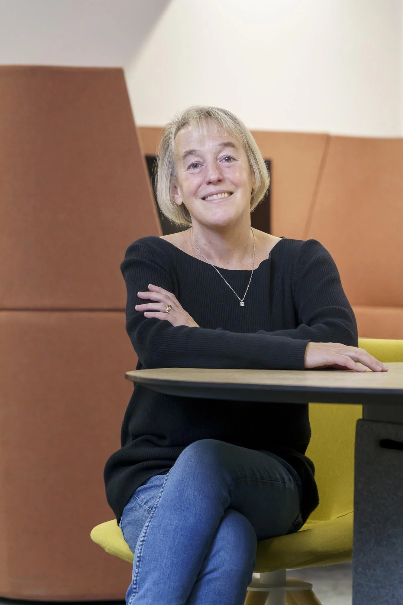 A women in her fifties sitting at a desk wearing a black jumper, blue jeans, and a necklace, with short blonde hair smiling as she shares how changing the food she eats improved her menopausal symptoms.