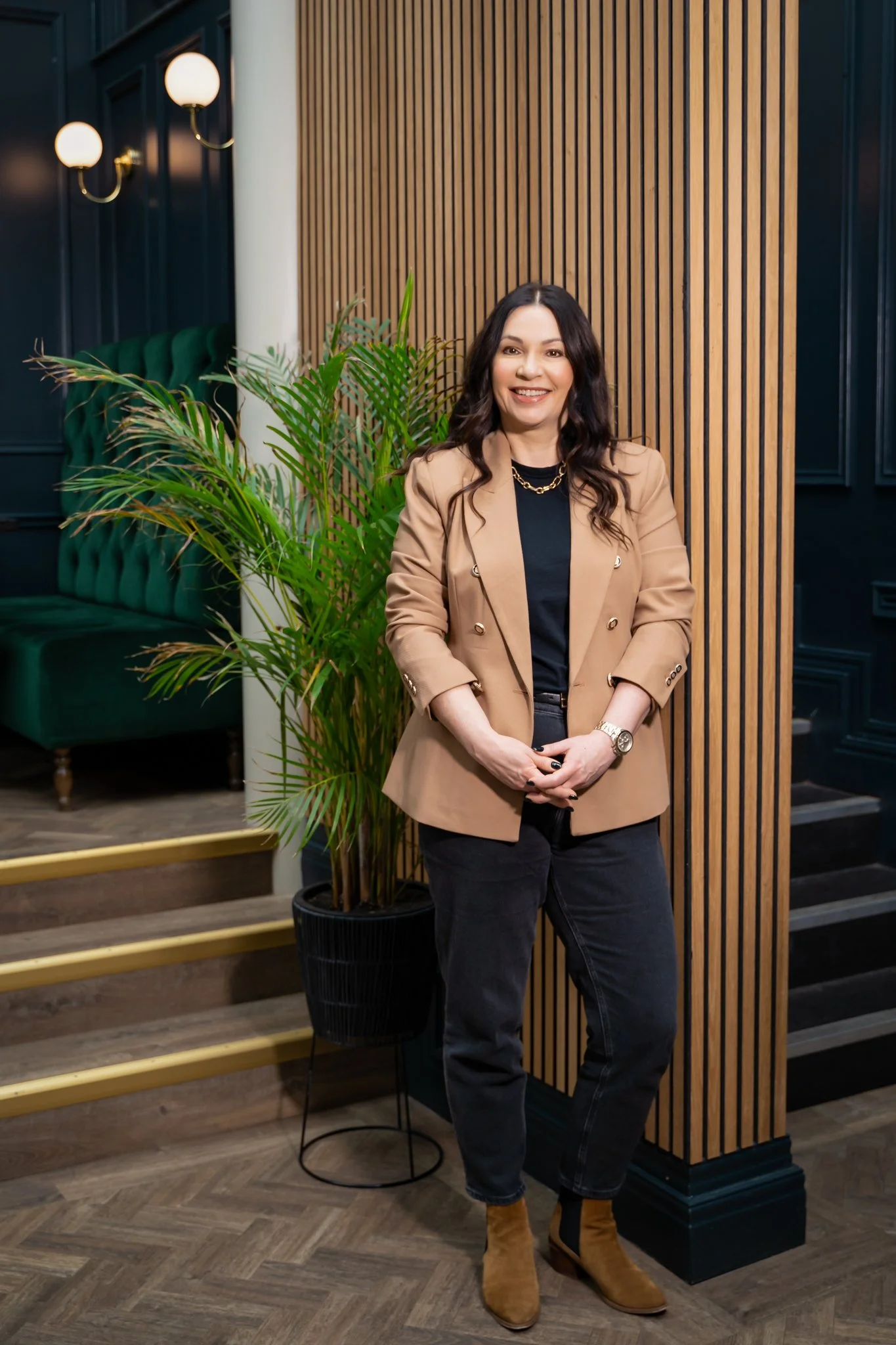 Nicola Farndell wearing black jeans, tan boots and a tan jacket with gold buttons, standing in front of a green plant and a wood panelled wall.