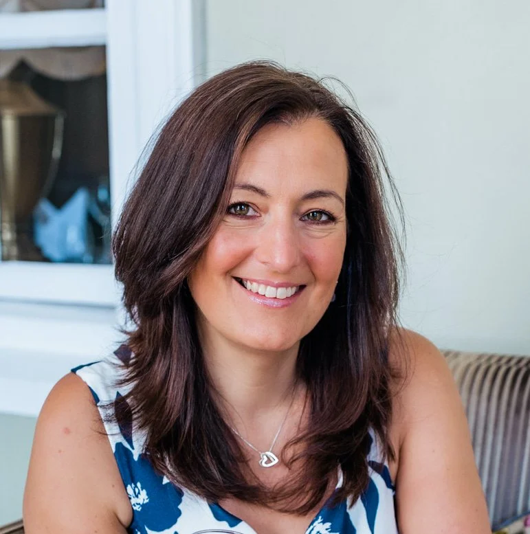 A midlife woman with dark brown hair over to one side and hanging over her chest wearing a silver heart necklace and a blue flowered top, smiling as she shares her menopause exhaustion and how she navigated it with tools to support strong wellbeing.