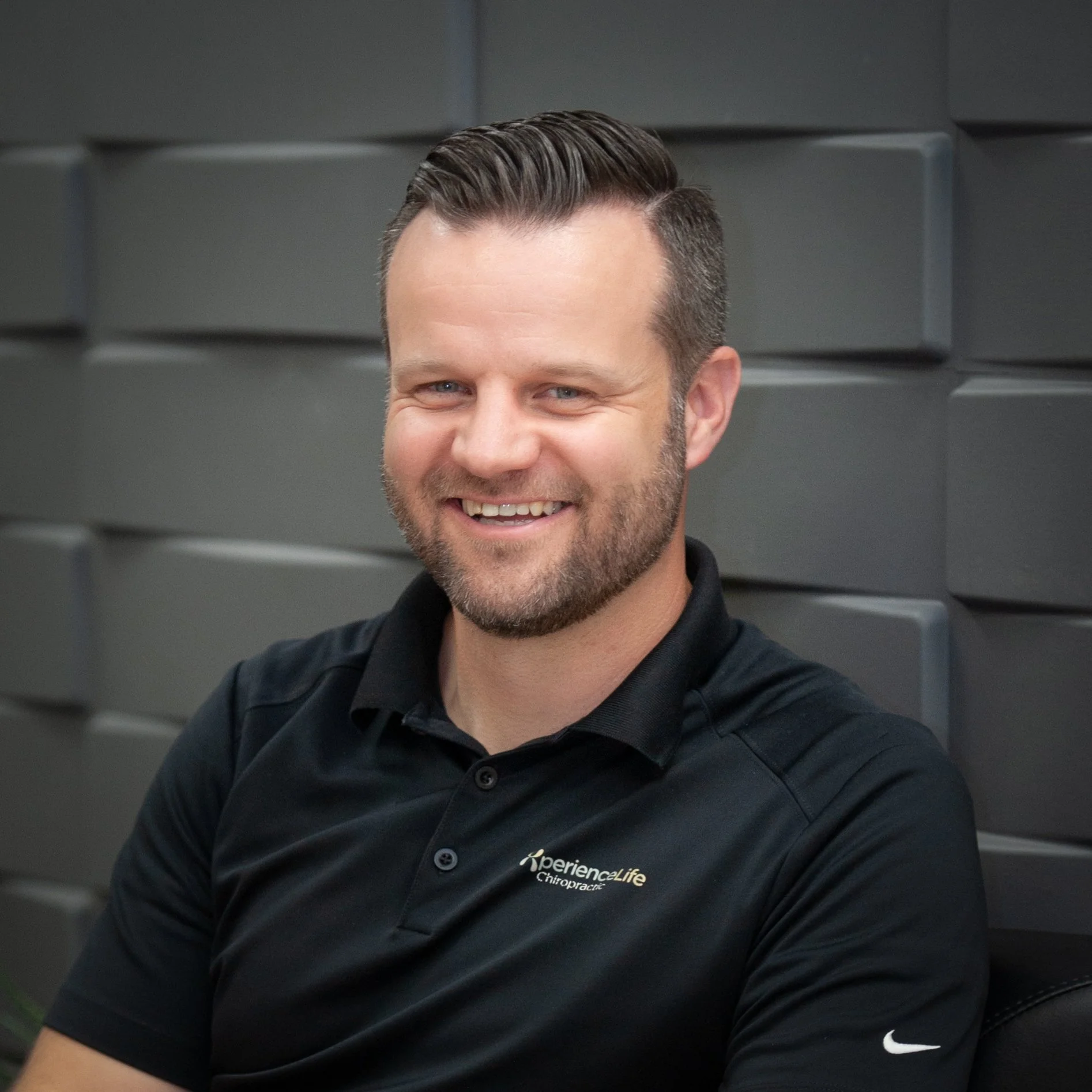 Dr Brett Hill, chiropractor and posture specialist, wearing a black polo top with light brown hair and a beard smiling as he shares the importance of posture for stress, energy, and brain fog in midlife.