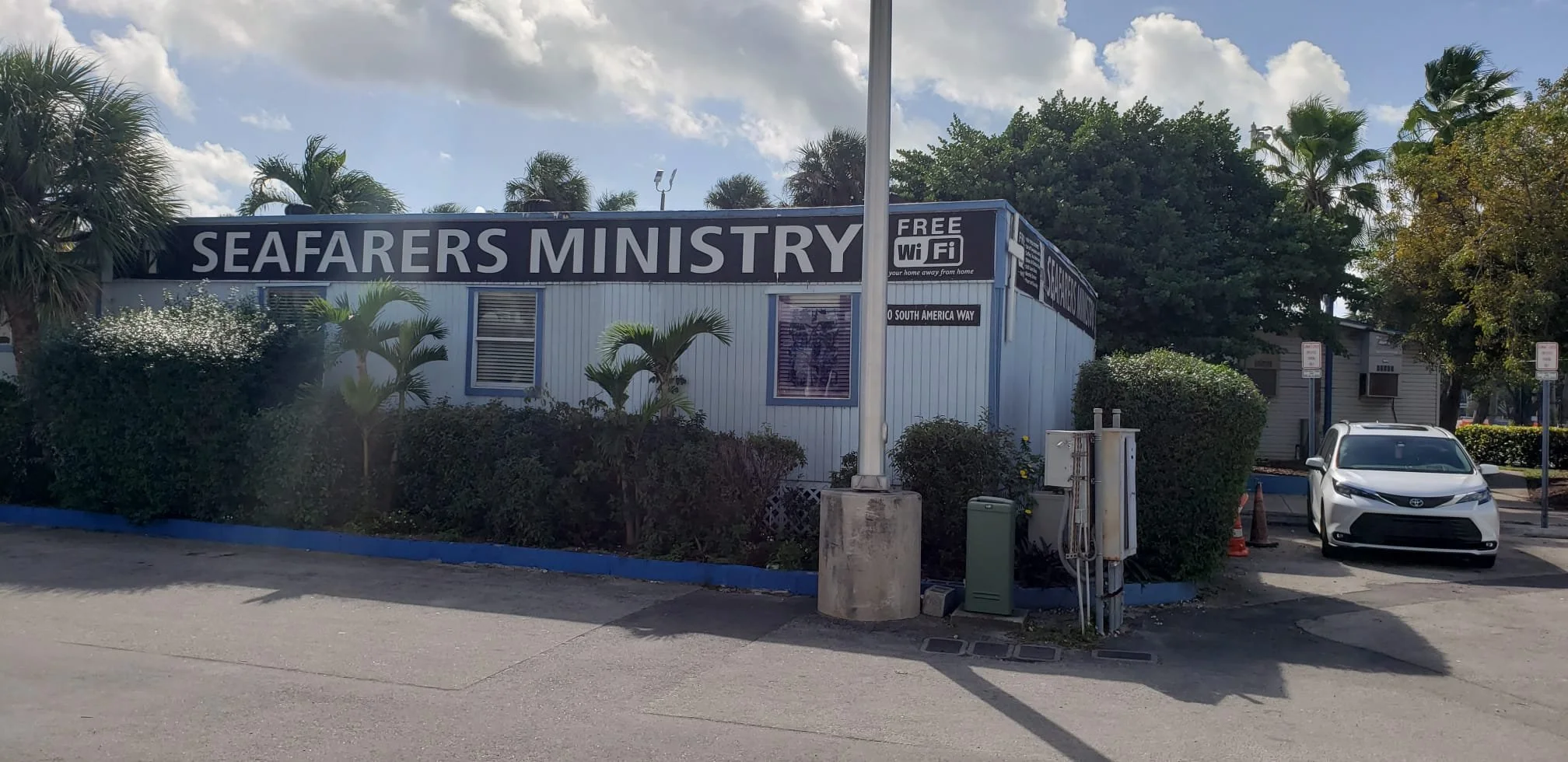 Exterior of a building with a sign that reads 'Seafarers Ministry', surrounded by trees and bushes, with a white car parked nearby and a sign indicating free Wi-Fi.