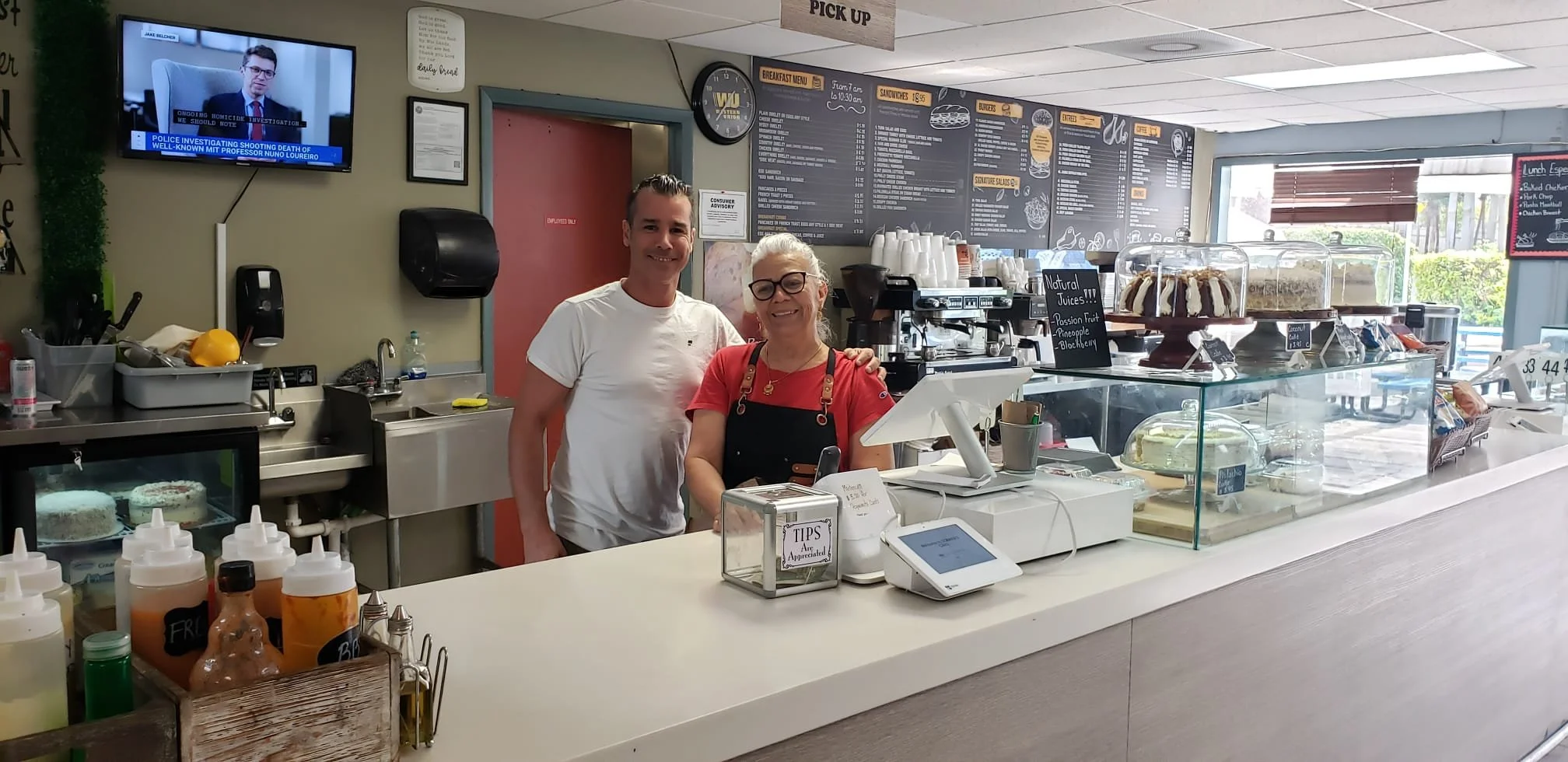 A man and a woman behind the counter of a bakery or cafe, smiling at the camera. The counter has a glass display case with cakes and baked goods, and various items like sauces and condiments in bottles. The background shows a menu board, a clock, and a TV screen. The woman is wearing an apron and the man is dressed casually.