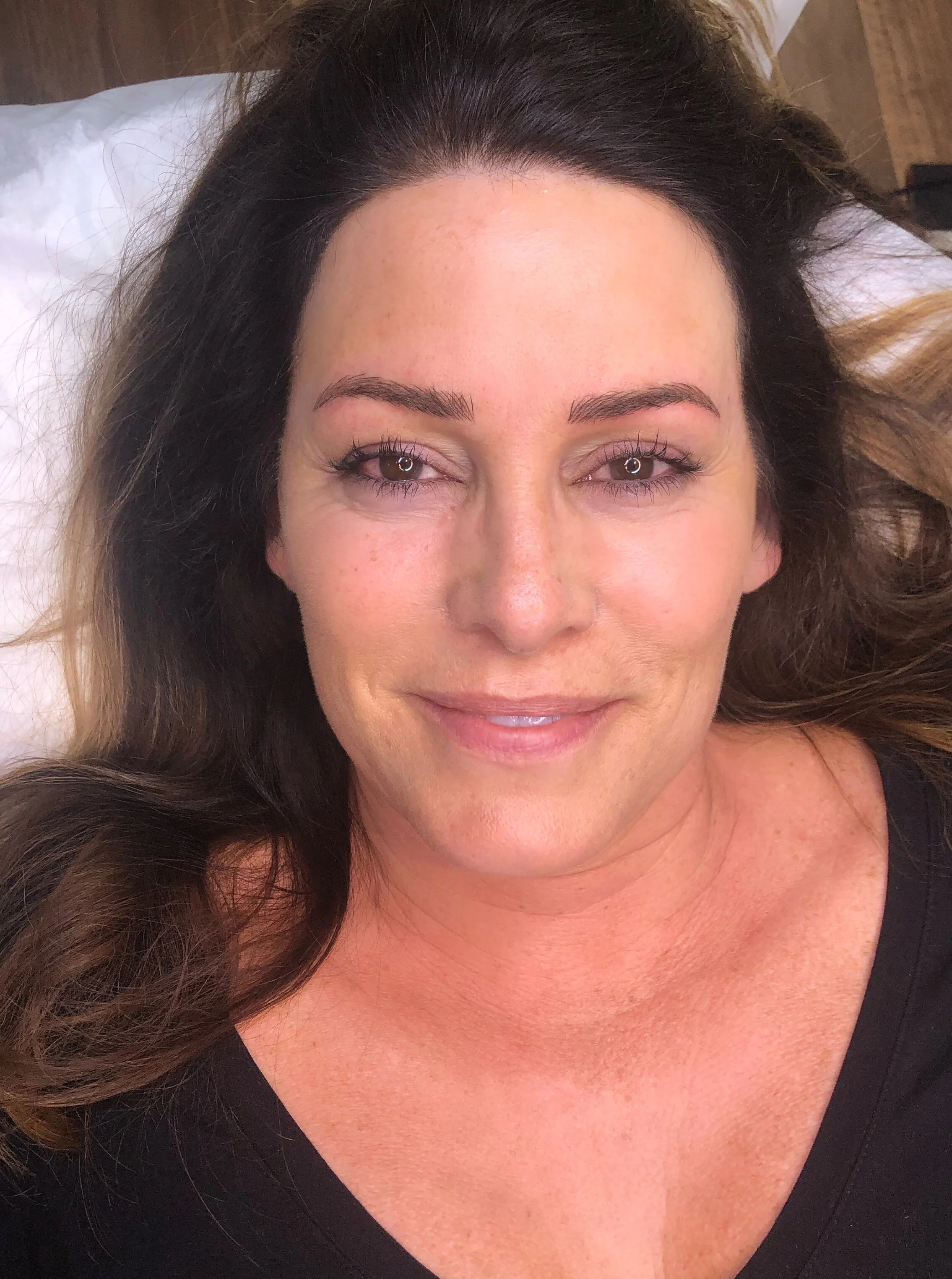 A woman with long brown hair and light skin lying on a bed, smiling at the camera.
