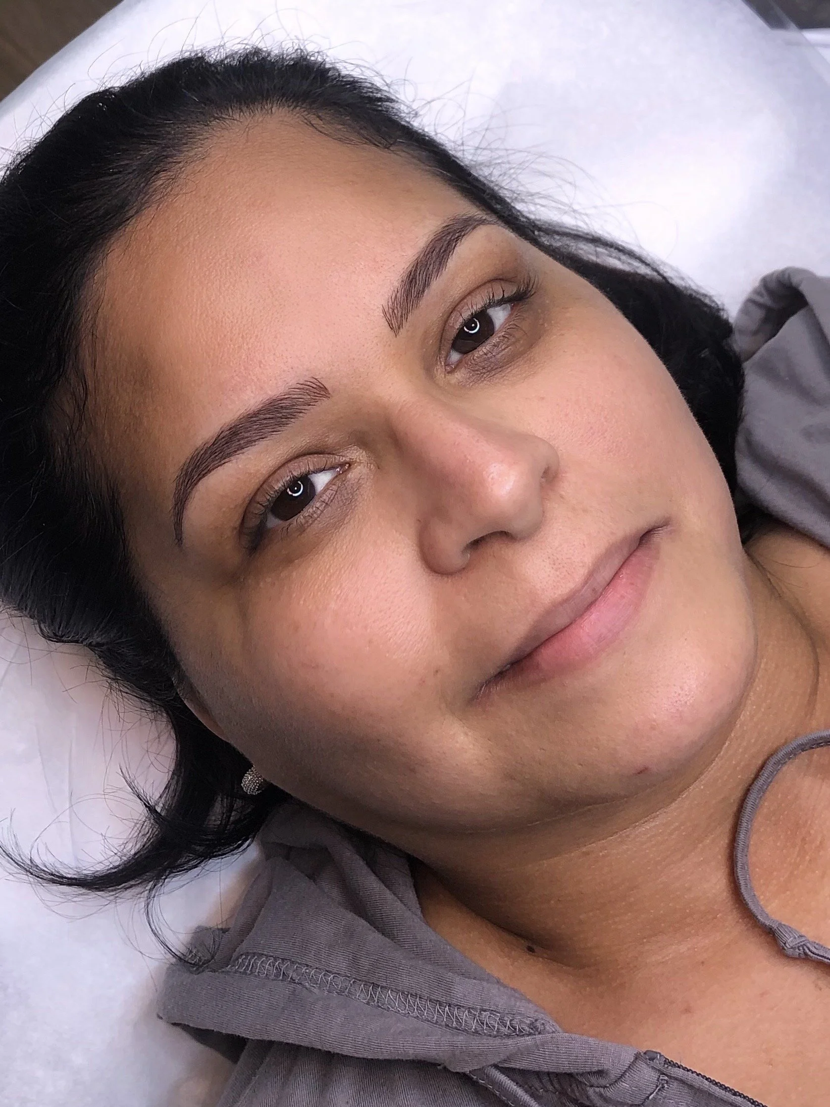 A close-up of a woman with dark hair and well-groomed eyebrows, lying down with her head on a pillow, looking at the camera.