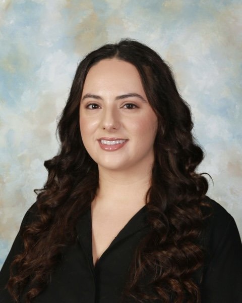 Portrait of a smiling young woman with long dark brown hair wearing a black blouse against a light blue with clouds background