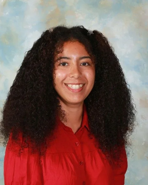 Portrait of a smiling young woman with wavy dark brown hair wearing a red collared blouse against a light blue with clouds background