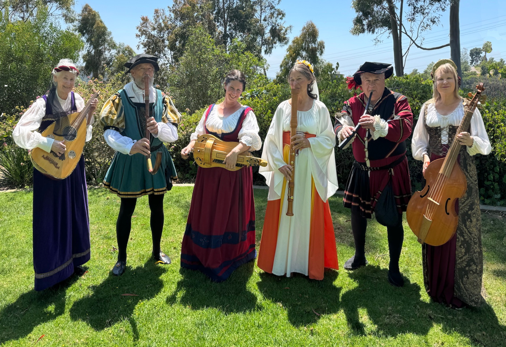 An outdoor picture of six performers standing on a grassy lawn in a line dressed in period costumes from feudal times with old fashioned period musical instruments.