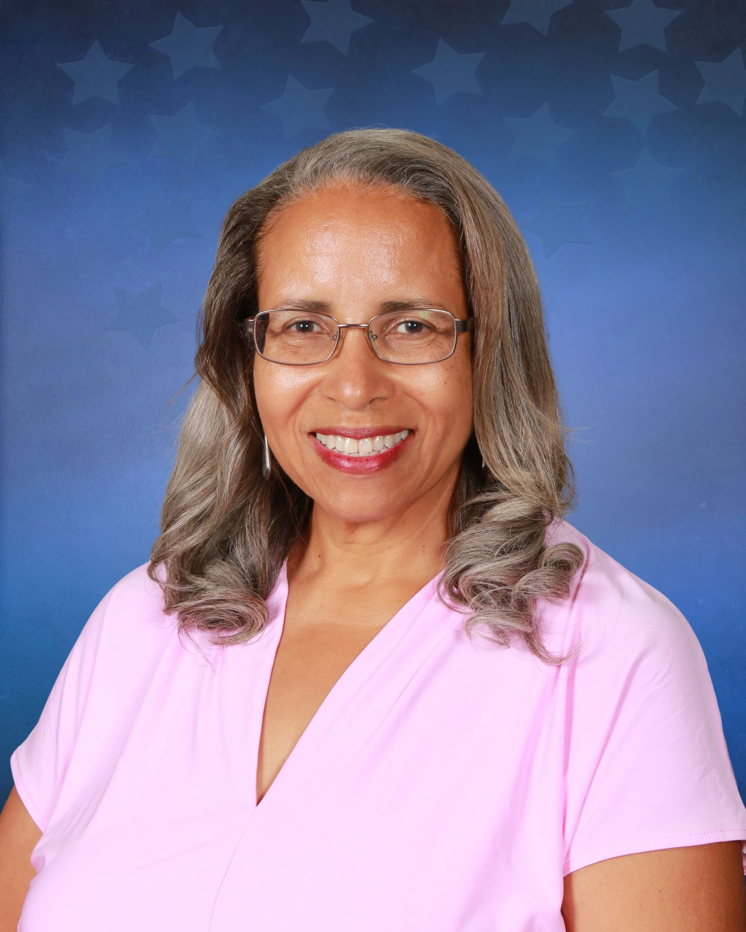 Portrait of a smiling woman with long brown hair, wearing hoop earrings and a pink v-neck blouse against a blue background