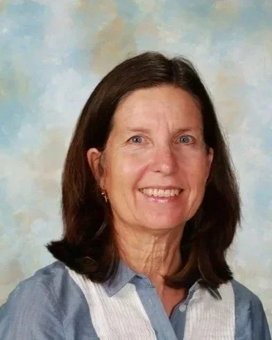 Portrait of a smiling woman with shoulder length brown hair, wearing a light blue collared shirt with white trim against a light blue with clouds background