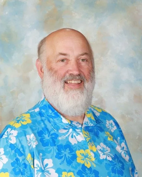 Portrait of a smiling man with a full white beard wearing a patterned floral blue, yellow and white Hawaiian collard button down shirt, against a patterned light blue with clouds background