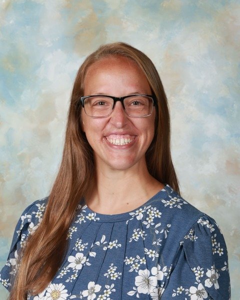 Portrait of a smiling woman wearing glasses with long dark blonde hair wearing a floral patterned blue and white blouse against a patterned light blue with clouds background