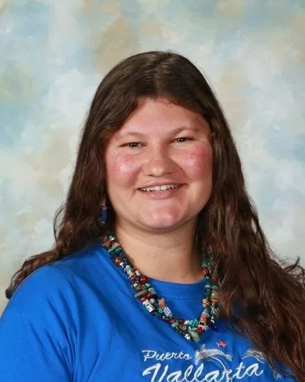 Portrait of a smiling woman with long brown hair wearing a royal blue shirt and a multi-strand beaded necklace against light blue and swirly clouds background