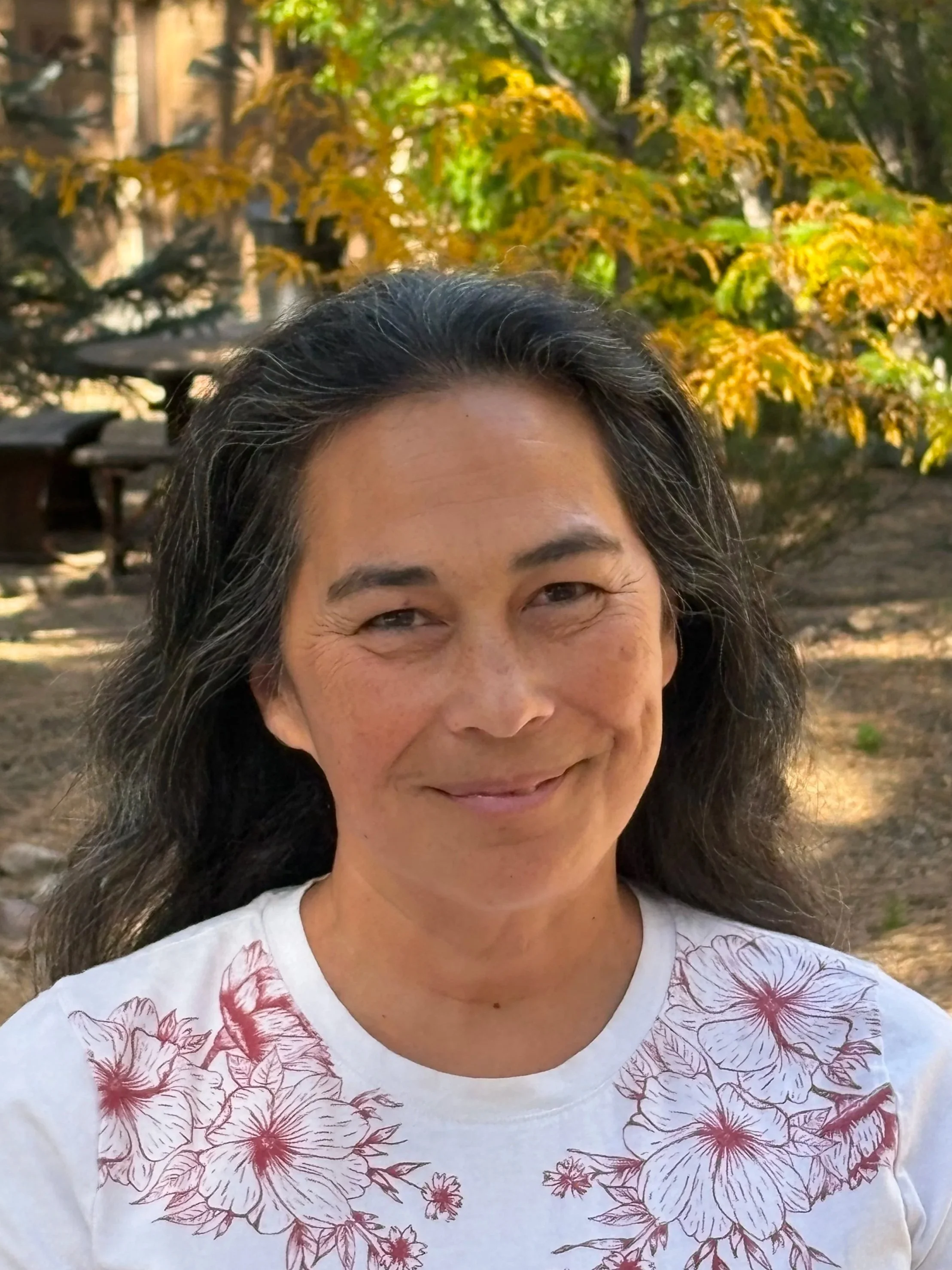 Head shot of a smiling woman with long brown hair wearing a white crew neck shirt with a pink floral sketch pattern outdoors with greenery behind her
