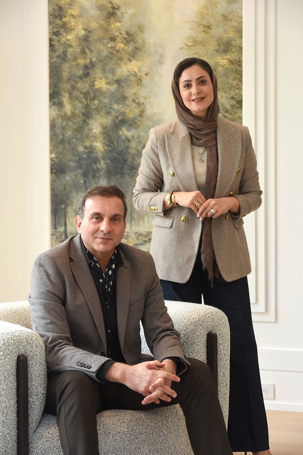 A woman in a beige blazer and hijab standing beside a seated man in a grey blazer, in a room with a large landscape painting in the background.