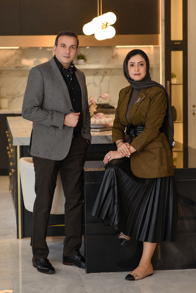A man in a gray blazer and dark pants standing next to a woman in a green jacket, black pleated skirt, and a dark scarf in a modern kitchen.