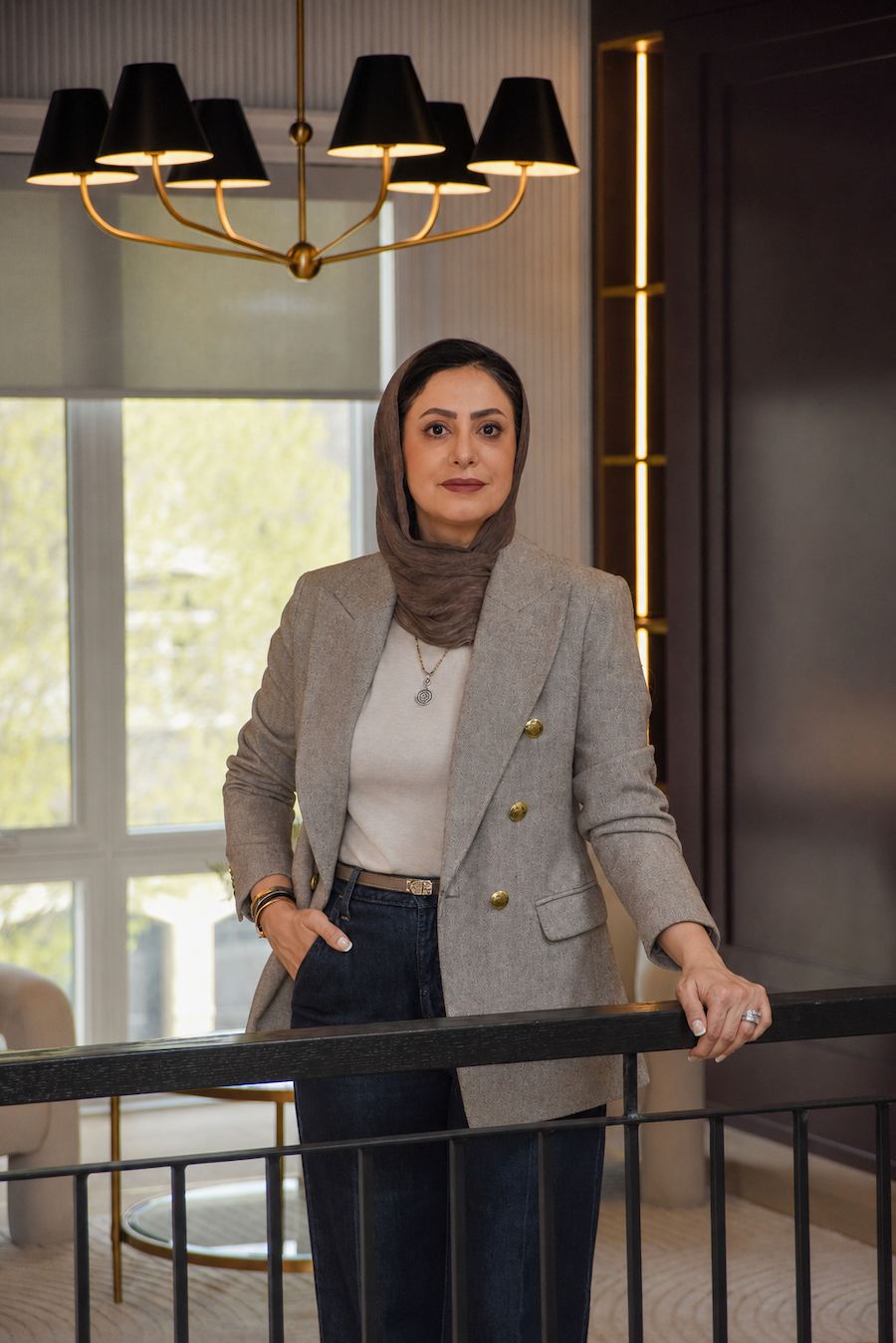 A woman wearing a beige blazer and dark jeans, with a headscarf, stands indoors near a black railing. She has short dark hair, makeup, and is accessorized with jewelry. There is a modern black and gold chandelier overhead and a window in the background with a view of trees outside.