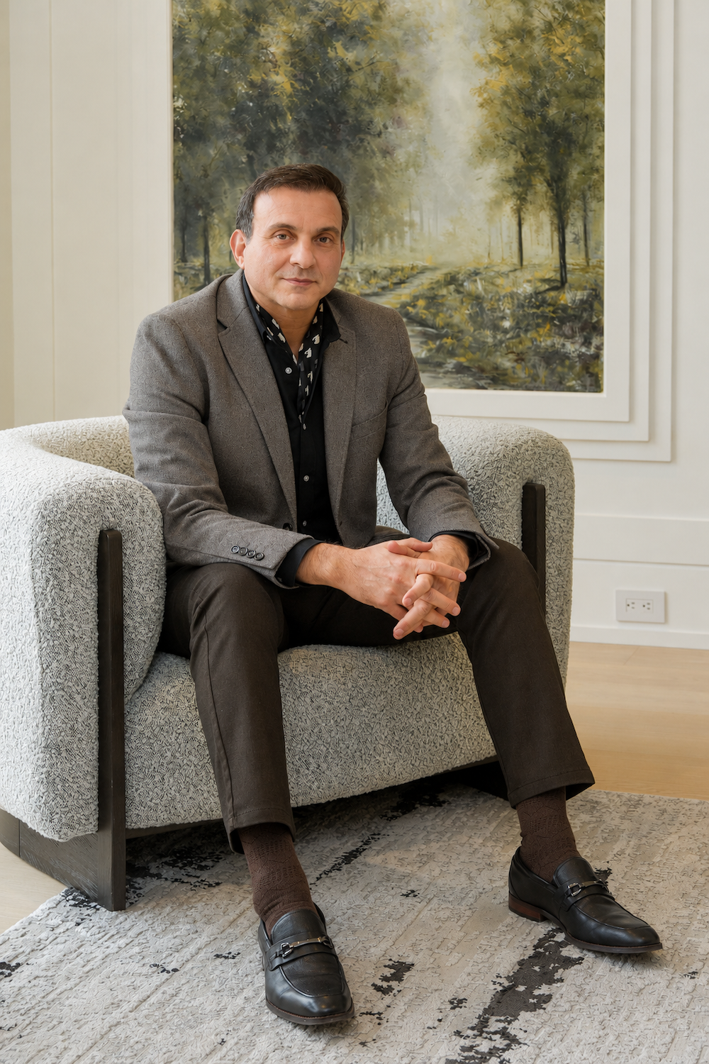 A man in a gray blazer and black shirt sitting on a light-colored sofa in a room with a large landscape painting of a forest trail on the wall behind him.