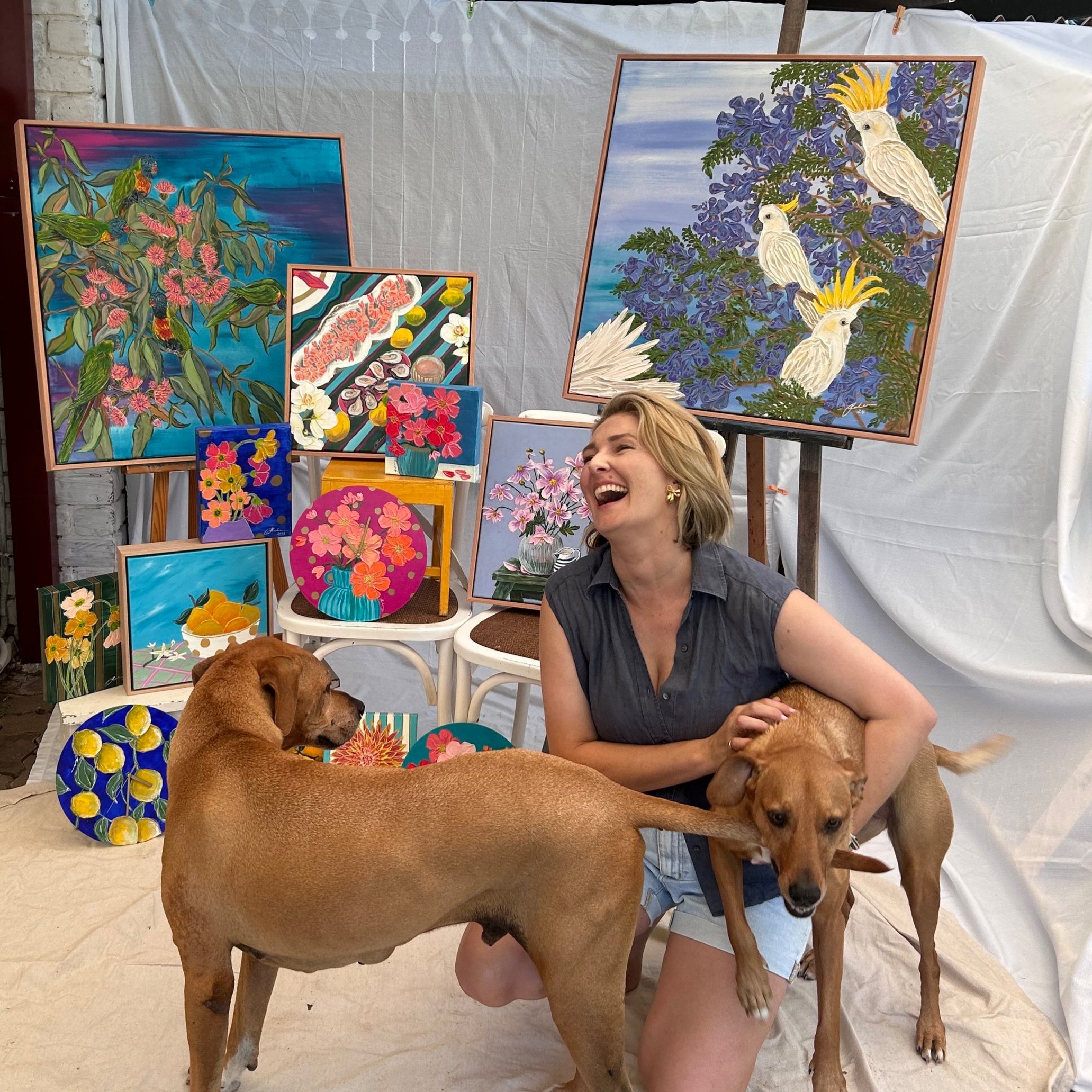 Australian artist Pippy Sheehan sits in front of her new collection of artworks created ahead of the Brisbane Etsy Made Local Market with her two dogs Cooper and Meg, all of them smiling