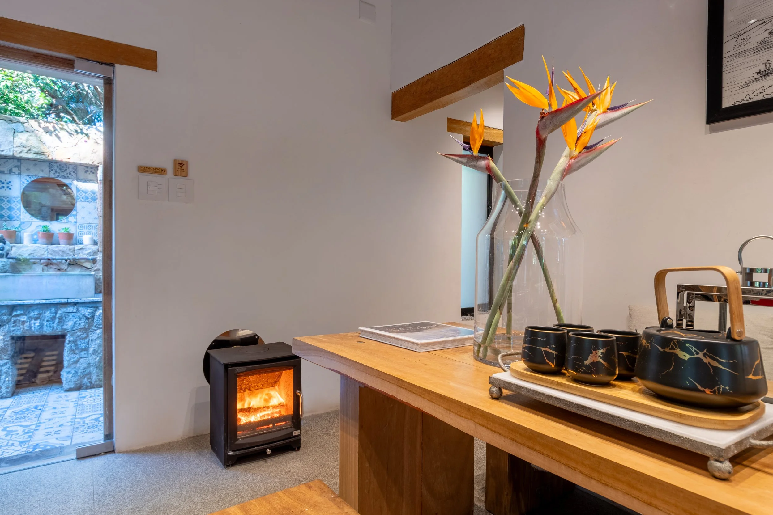 A wooden table with a decorative tray holding a black and gold teapot and cups, and a tall glass vase with tropical orange and purple flowers, in a cozy room with a small fireplace and a door leading outside.