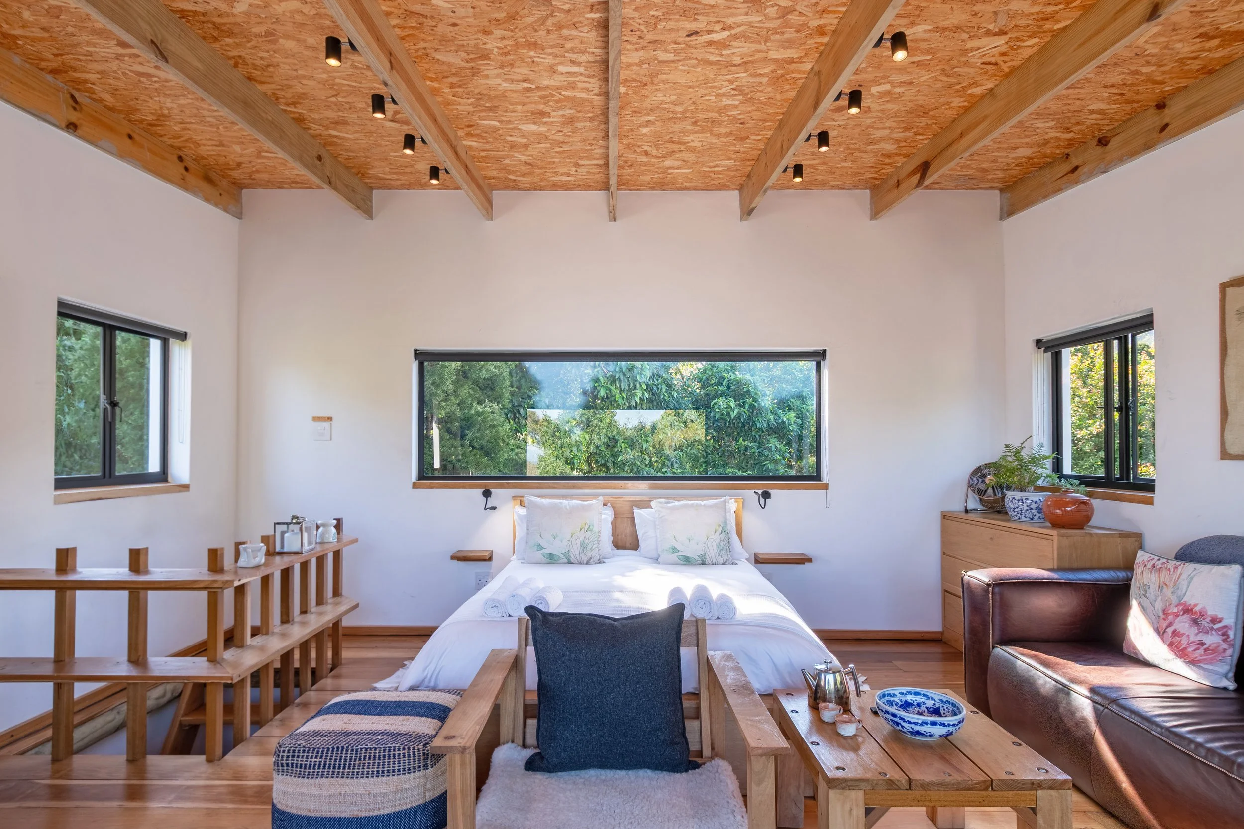 A cozy bedroom featuring a bed with white linens and decorative pillows, flanked by black reading lights, with large windows showing green trees outside. The ceiling has wooden beams and a textured wood finish. There are wooden furniture pieces, incl