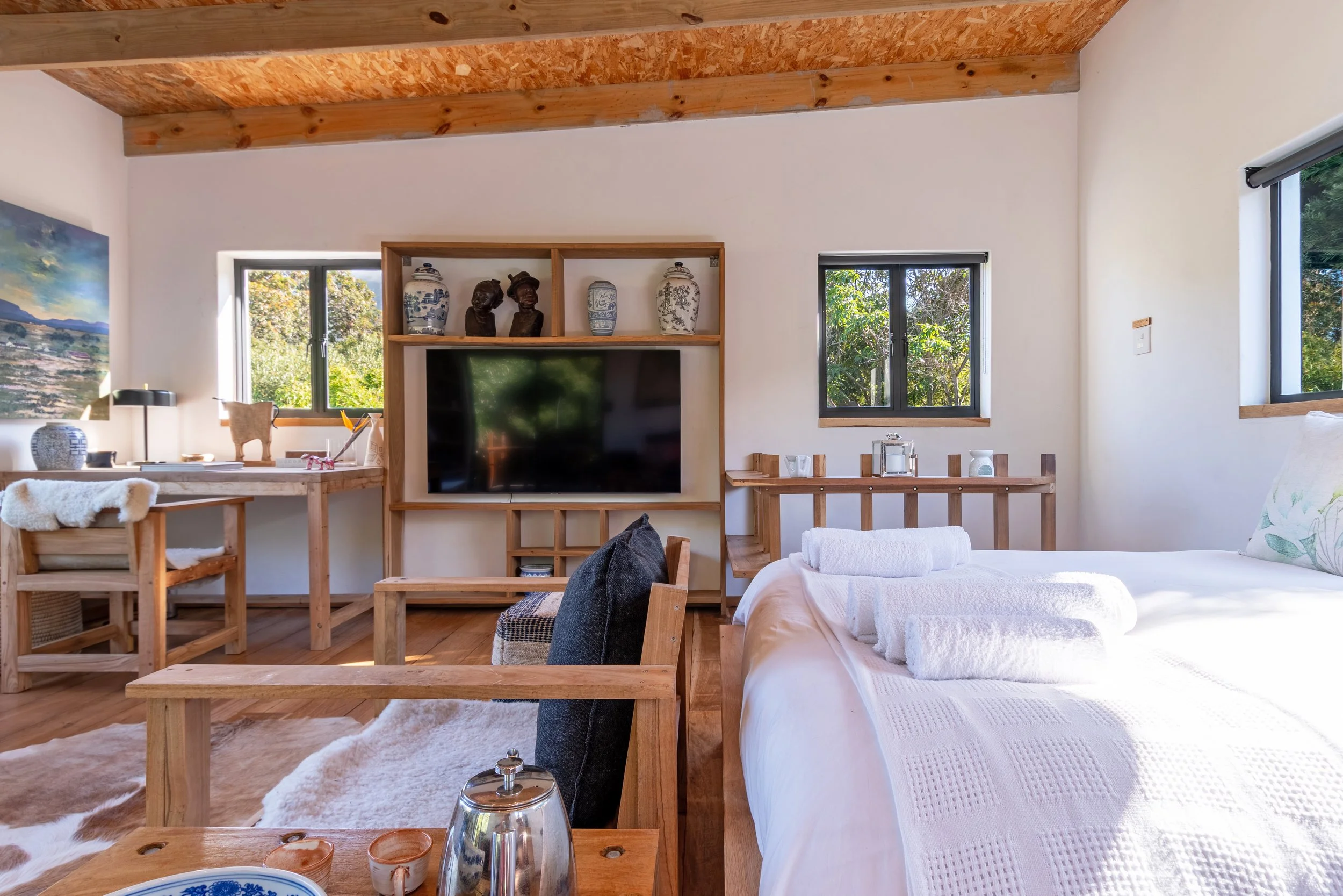 A cozy, well-lit bedroom with wooden beams, a white bed with rolled towels, a flat-screen TV on a wooden shelving unit, a desk with decorative items, and two windows showing greenery outside.