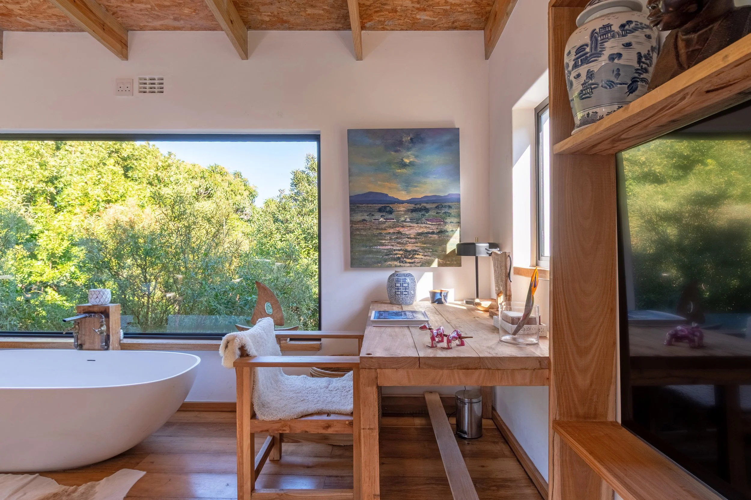 Interior of a modern living space with a white freestanding bathtub near a large window with greenery outside, a wooden desk with decor and a chair with a sheepskin throw, a blue and white vase, a landscape painting on the wall, and a wooden ceiling